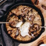 A black cast iron pan sits on top of a blue tea towel, on a wooden table. In the cut iron pan is a freshly baked chocolate caramel skillet cookie that has melty ice cream in the centre.