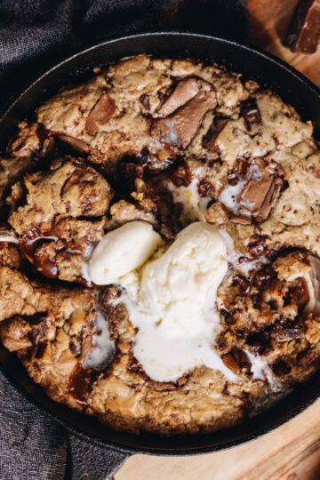 A black cast iron pan sits on top of a blue tea towel, on a wooden table. In the cut iron pan is a freshly baked chocolate caramel skillet cookie that has melty ice cream in the centre.