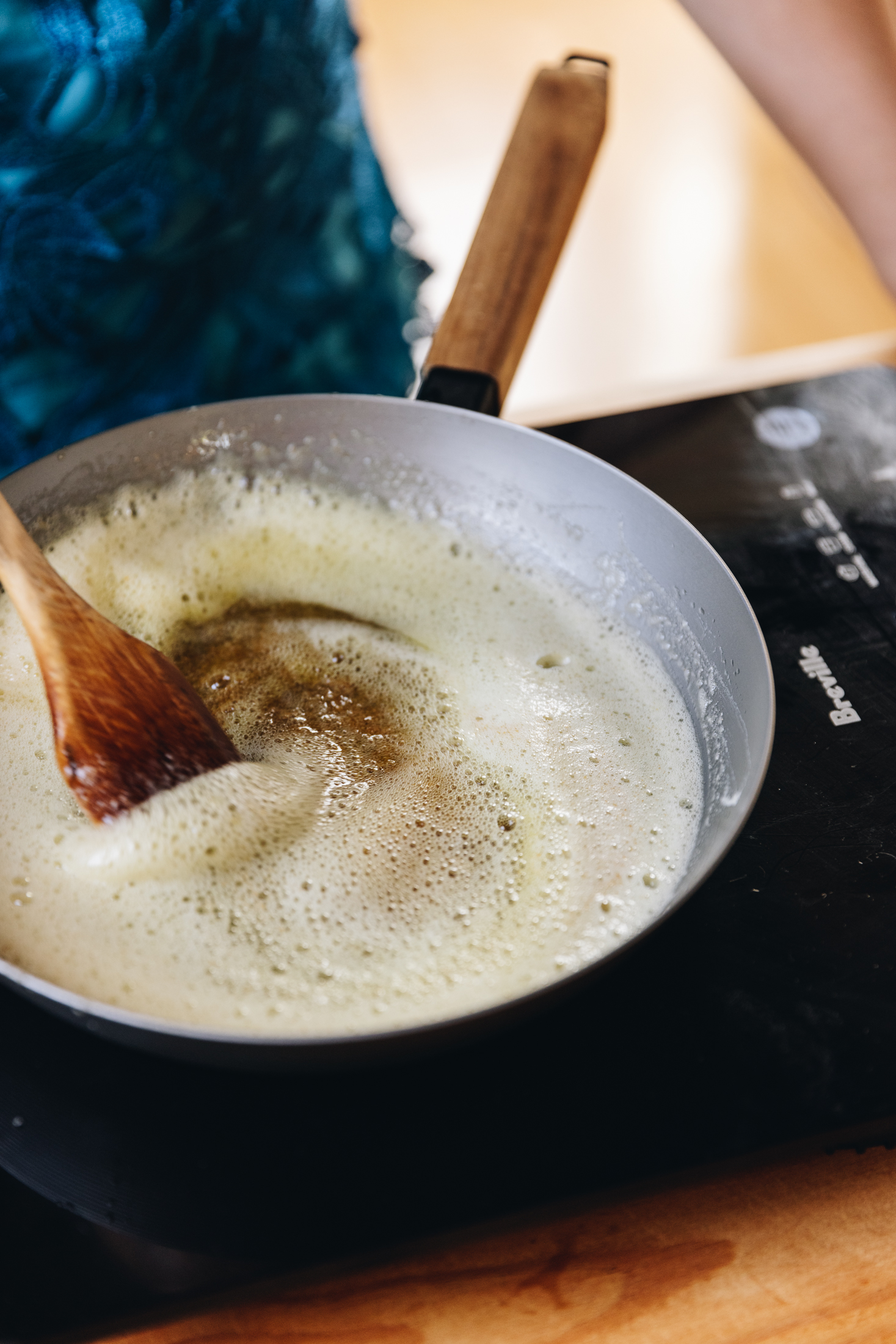A grey cast iron pan with a wooden handle sits on a black portable stove that sits on a wooden table. In the pan is foamy brown butter that is being stirred with a wooden spoon. 