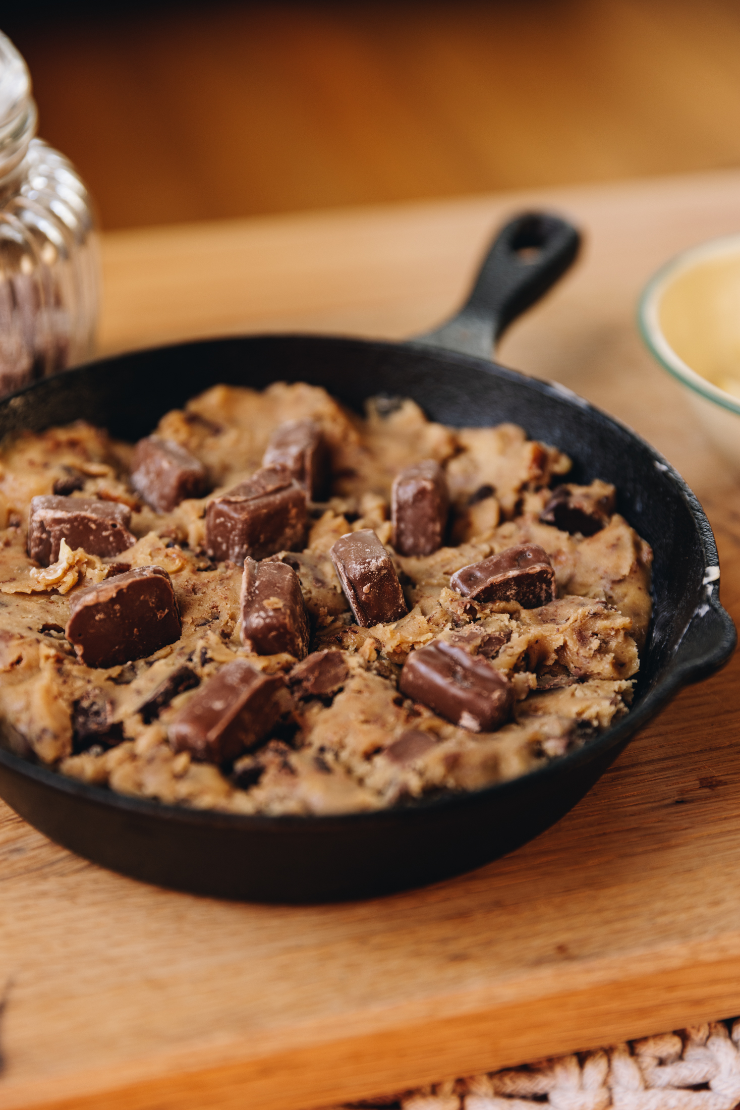 A black cast iron pan sits on a wooden board on a wooden table. In the pan is unbaked cookie dough that is studded with chocolate and chewy caramels. A glass jar and a small yellow bowl are on either side of the pan. 