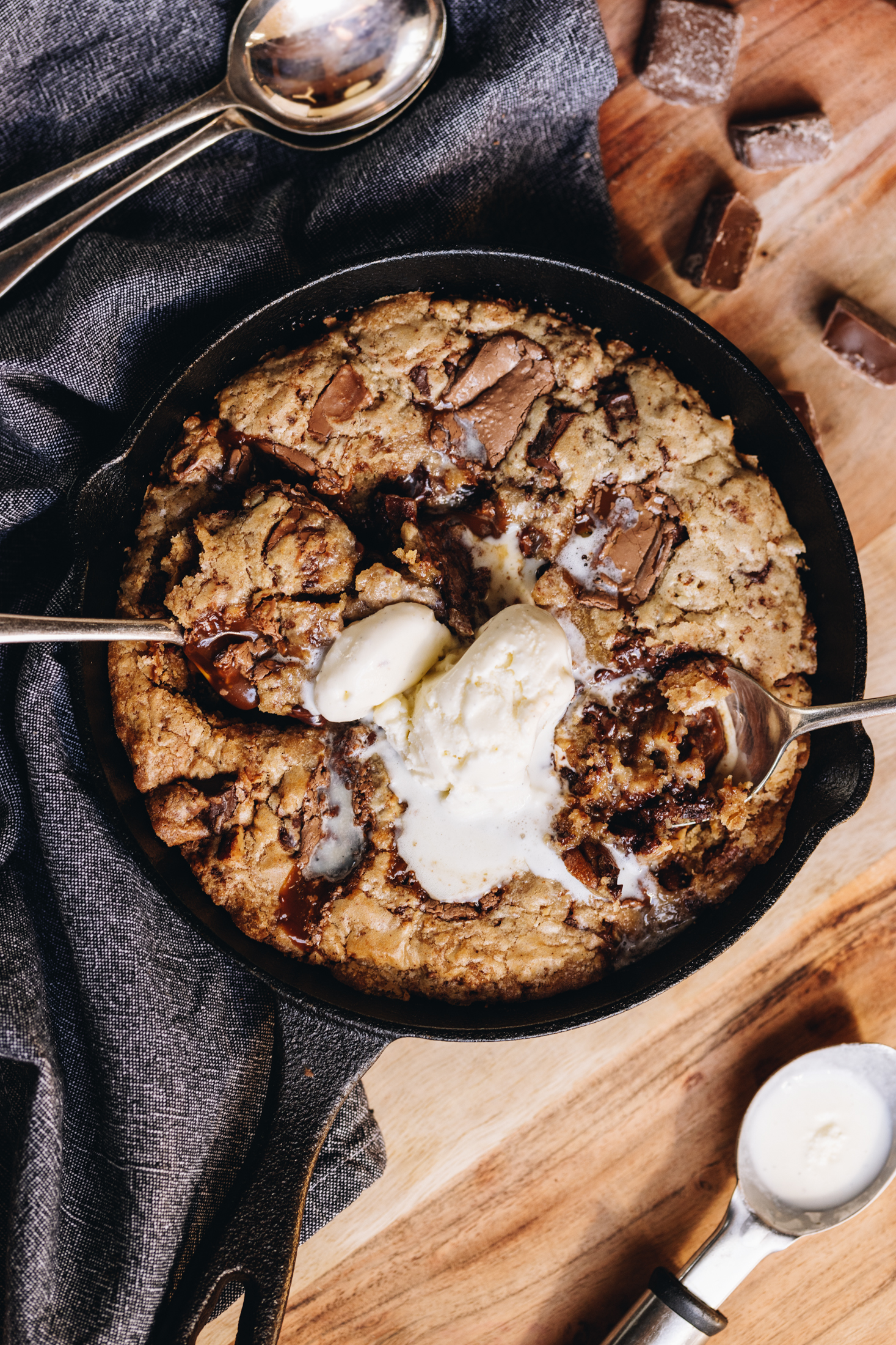 A black cast iron pan sits on top of a blue tea towel, on a wooden table. In the cut iron pan is a freshly baked chocolate caramel skillet cookie that has melty ice cream in the centre.