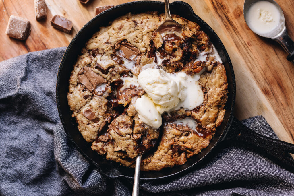 A black cast iron pan sits on top of a blue tea towel, on a wooden table. In the cut iron pan is a freshly baked chocolate caramel skillet cookie that has melty ice cream in the centre.