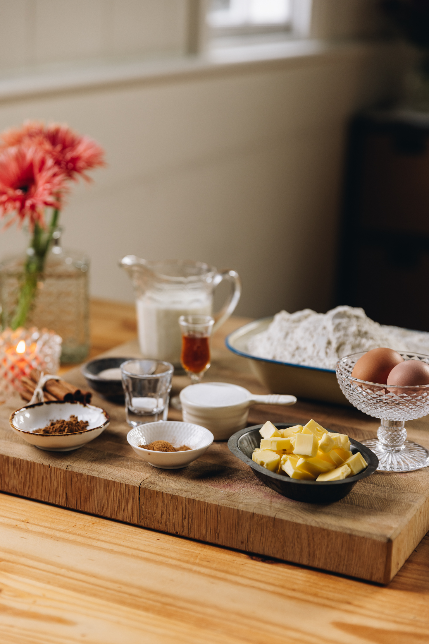 A wooden board sits on a wooden table. Vintage bowls and jars have ingredients in them. There is butter, spices, vanilla, eggs, flour and milk all in view. A glass with an orange candle burns next to the board and a glass vase with peach flowers in it is next to the candle.