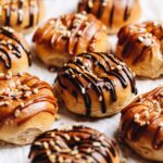 A tray of gooey chocolate baked doughnuts are on white baking paper. Each one is drizzled with either chocolate or caramel sauce and sprinkled with chocolate and caramel sprinkles.