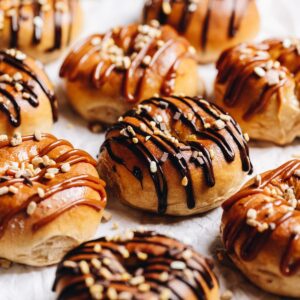 A tray of gooey chocolate baked doughnuts are on white baking paper. Each one is drizzled with either chocolate or caramel sauce and sprinkled with chocolate and caramel sprinkles.