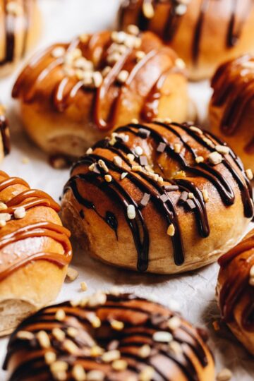 A tray of gooey chocolate baked doughnuts are on white baking paper. Each one is drizzled with either chocolate or caramel sauce and sprinkled with chocolate and caramel sprinkles.