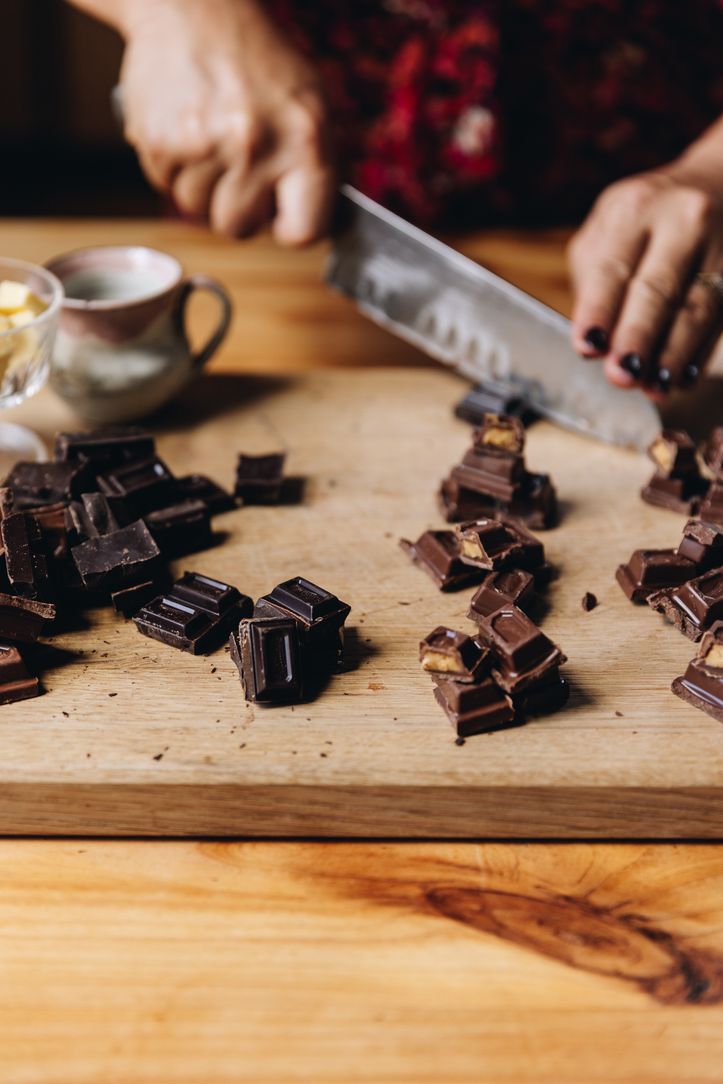 A wooden board sits on a wooden table. On the board is different types on chocolate that are in small piles. Naomi is cutting the chocolate in to pieces with a large silver knife.