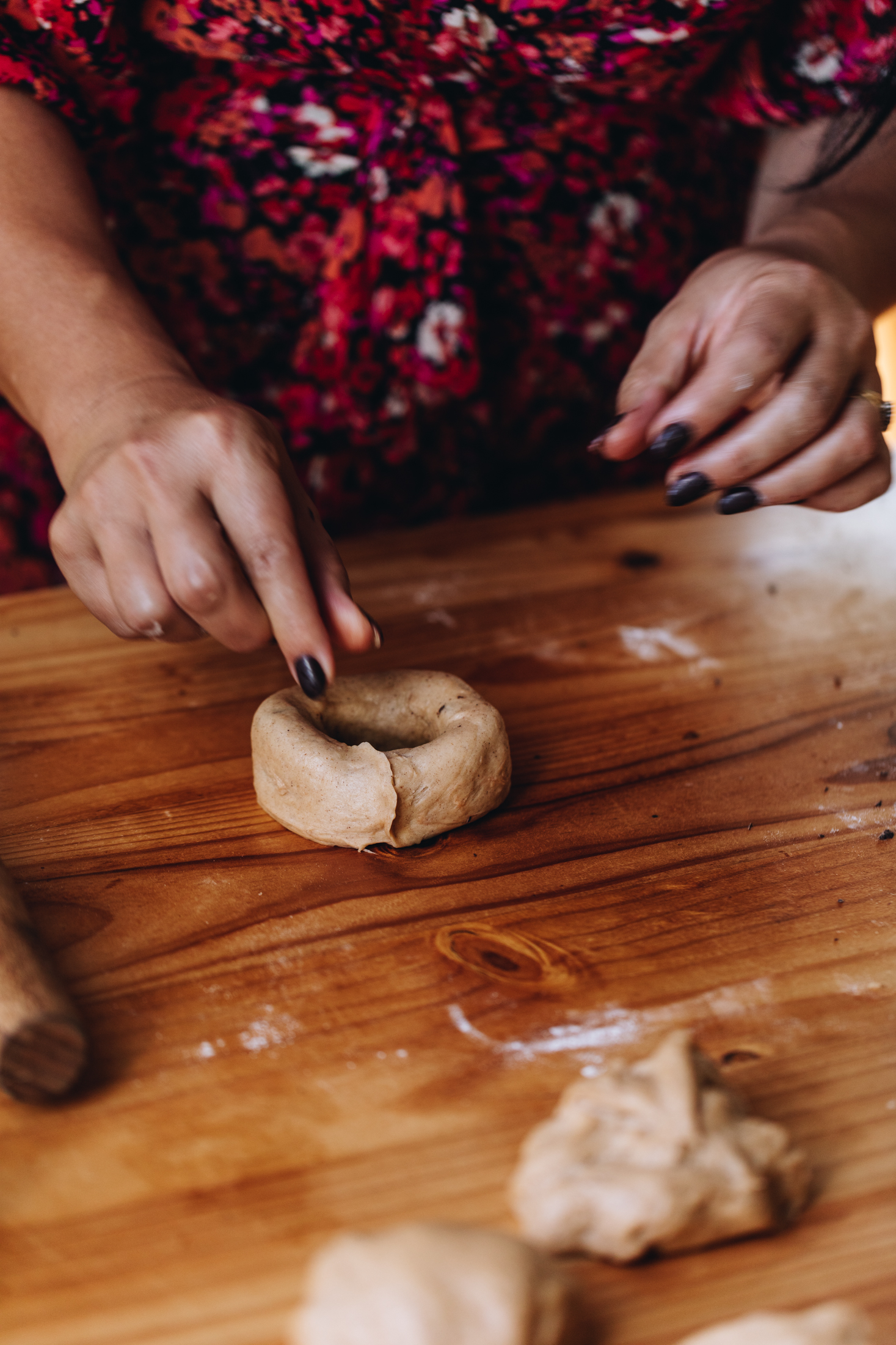 On a wooden table is a piece of dough that has been shaped in to a doughnut shape. Naomi is about to pinch the ends together. More blobs of dough at the front of shot.