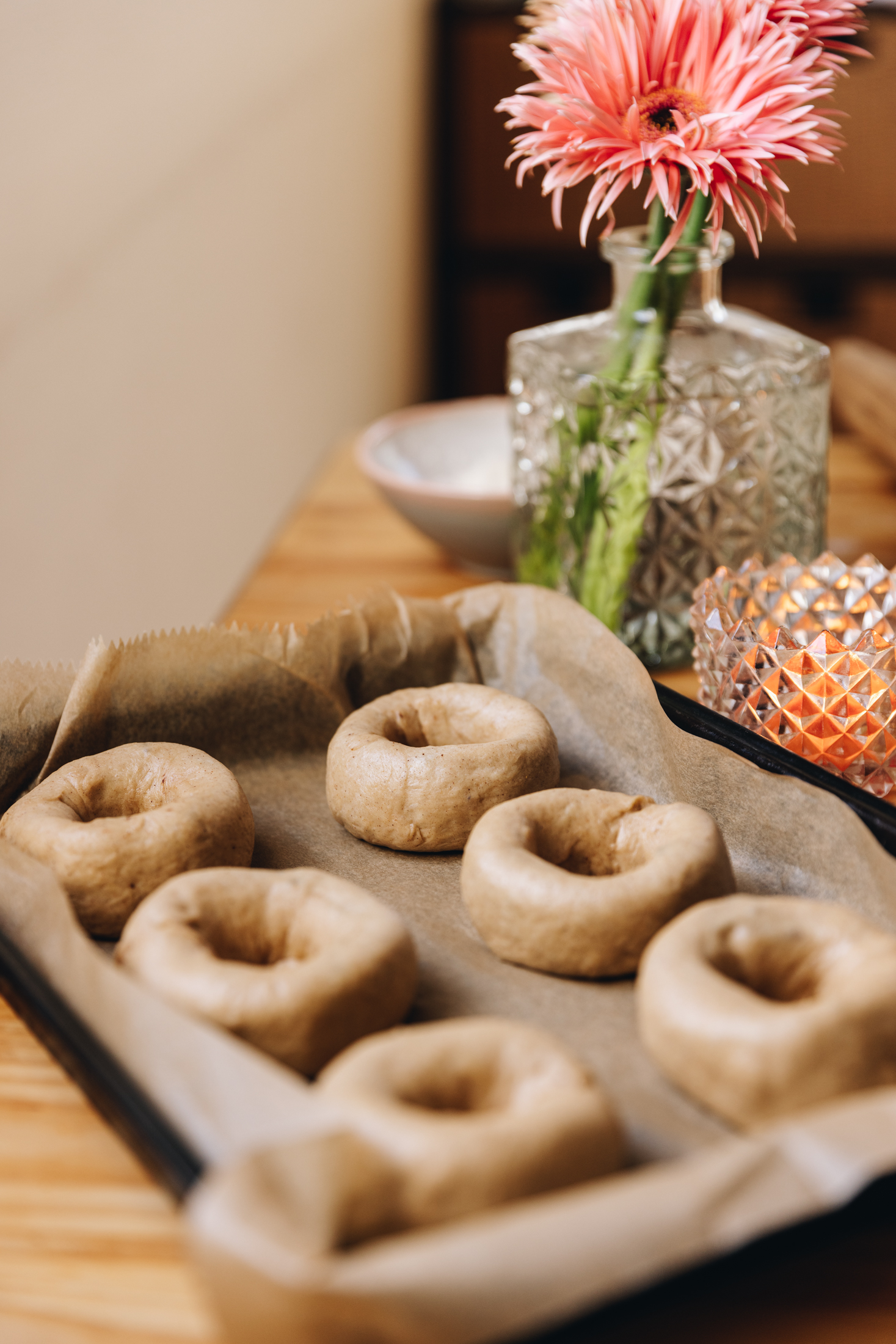 A black tray lined with brown baking paper sits on a wooden table. In the tray is six gooey chocolate baked doughnuts that are unbaked. Next to the tray is an orange candle in a glass jar and peach flowers in a vintage glass vase.