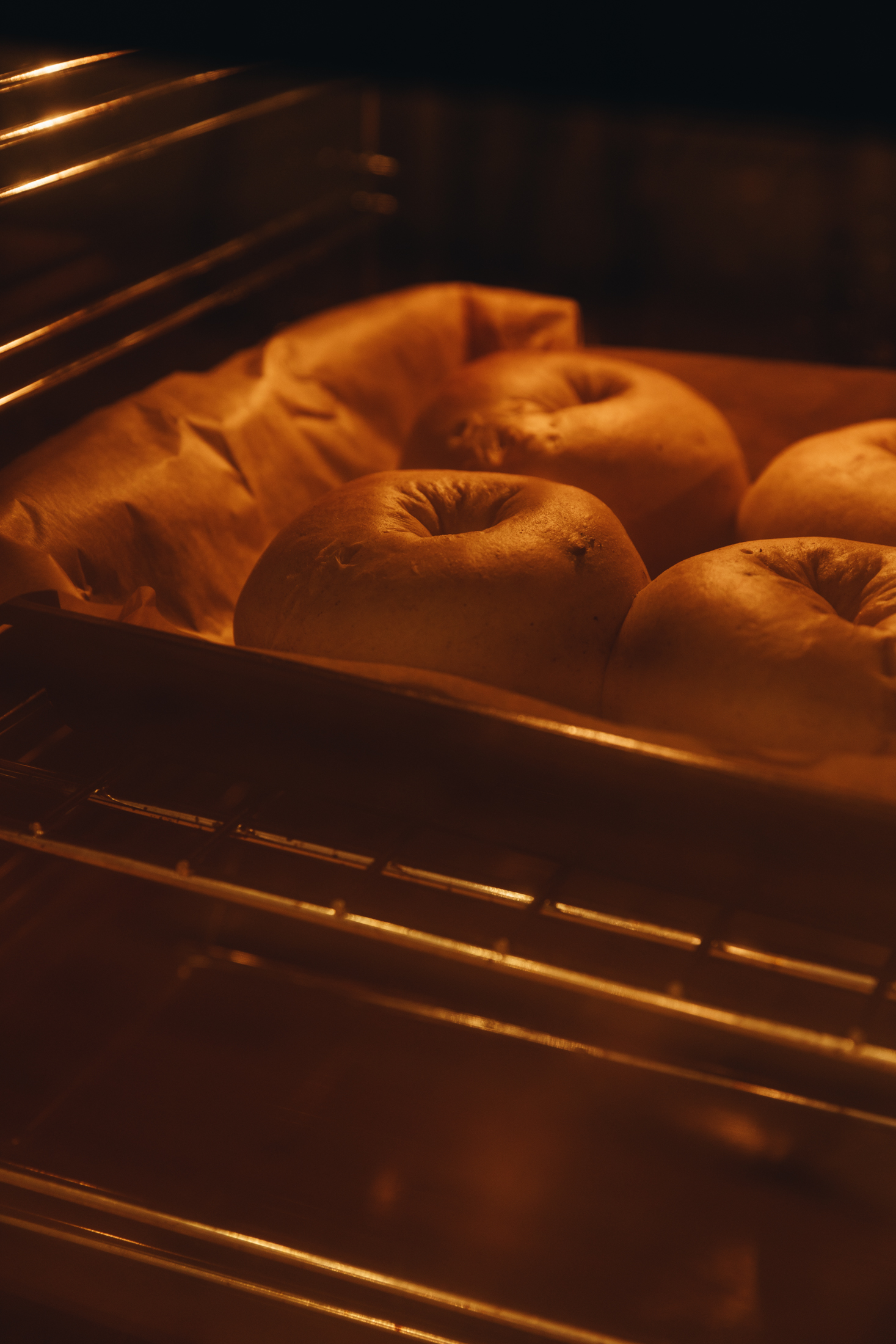 In an oven a tray lined with brown baking paper has baked doughnuts baking in it.