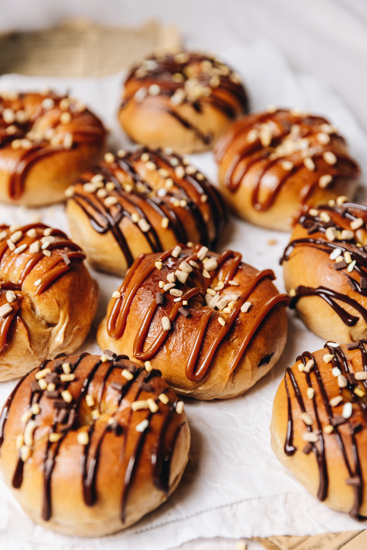 A tray of gooey chocolate baked doughnuts are on white baking paper. Each one is drizzled with either chocolate or caramel sauce and sprinkled with chocolate and caramel sprinkles.