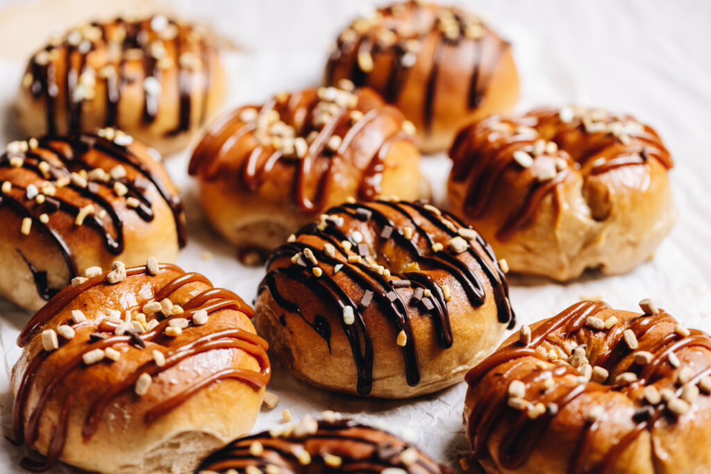 A tray of gooey chocolate baked doughnuts are on white baking paper. Each one is drizzled with either chocolate or caramel sauce and sprinkled with chocolate and caramel sprinkles.