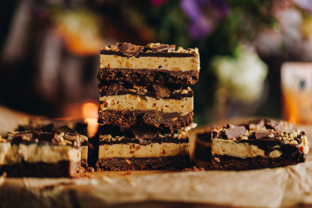 On a wooden board, lined with brown baking paper is five pieces of peanut butter weetbix slice. One piece sits on the left nd one on the right side. In the centre is a stack of three slices. They all have a chocolate weetbix base, a peanut butter filling and chocolate on top. 