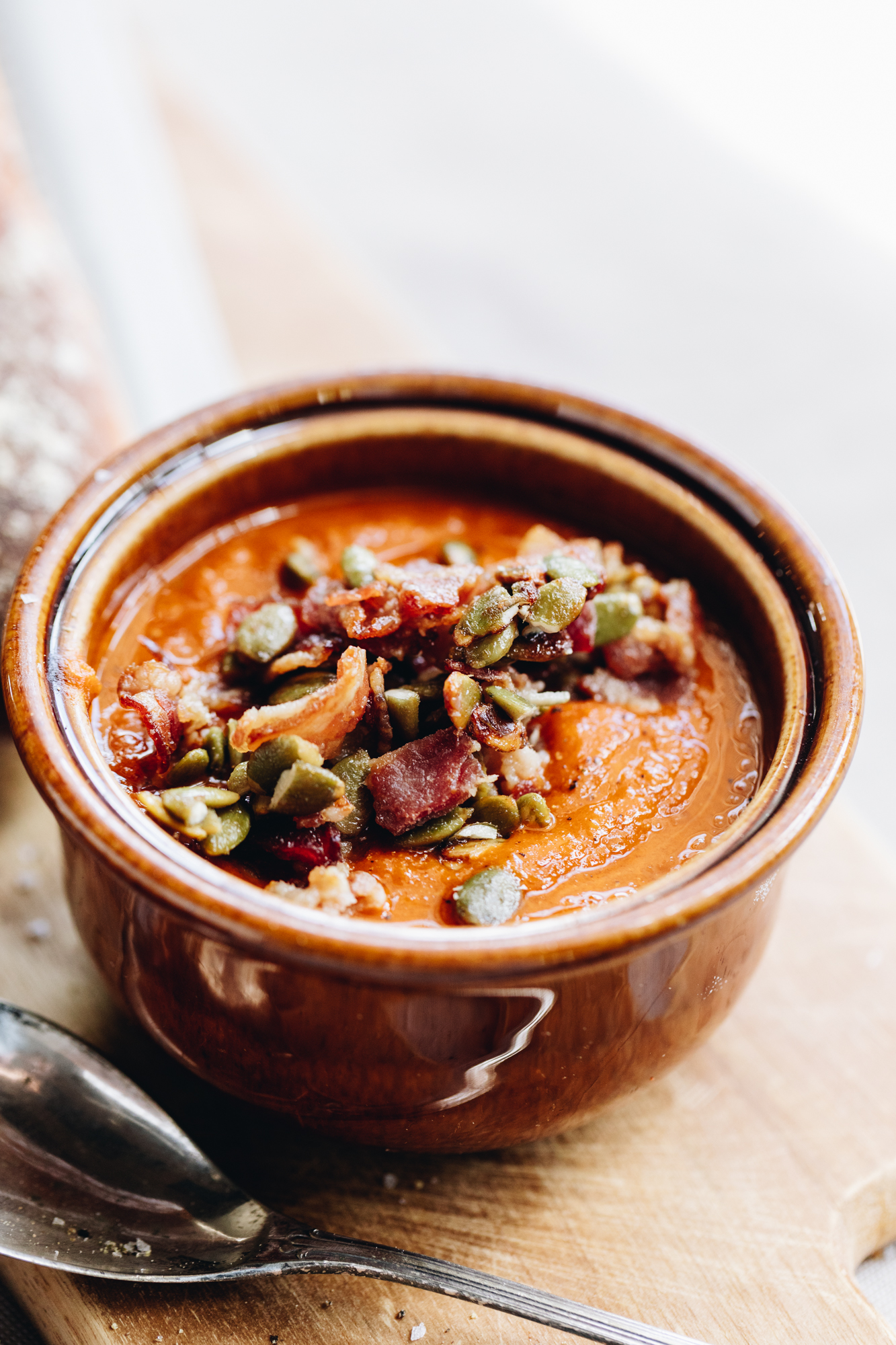 A brown ceramic bowl sits on a wooden board with a silver spoon next to it. In the bowl is easy roasted tomato soup. The soup is topped with bacon and pumpkin seeds.