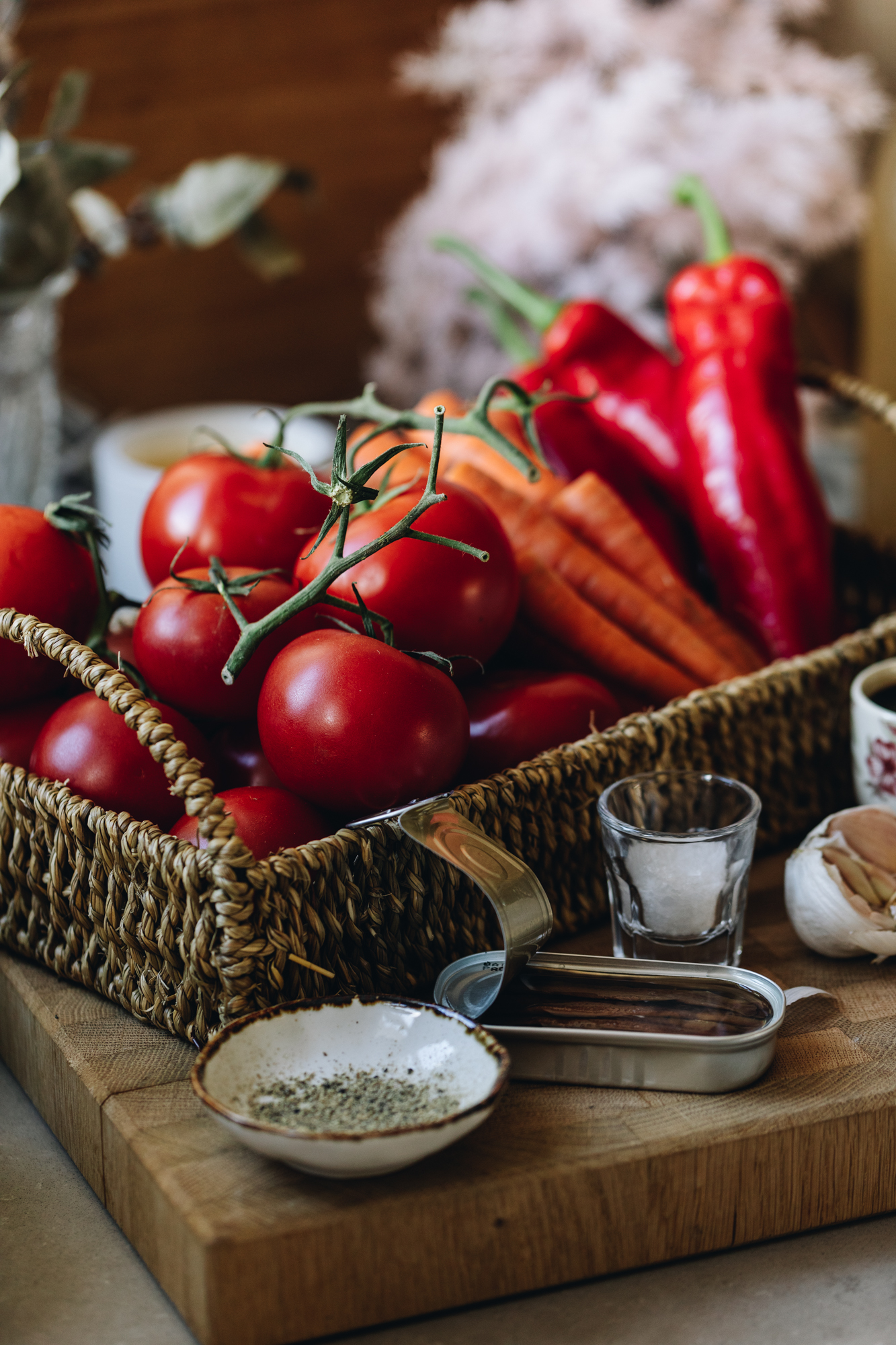 On a stone bench sits a wooden board. On the board is can basket with fresh tomatoes, carrots and capsicum in it. At the front of the board is pepper, anchovies, salt and garlic. 