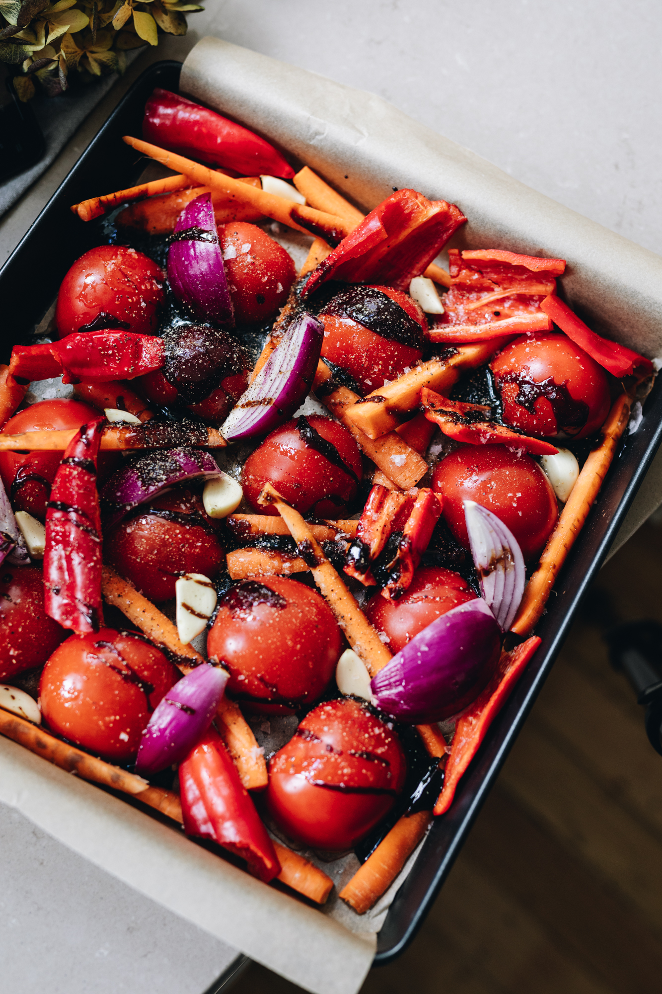 A roasting tray lined with baking paper sits on a stone bench. In the tray is tomatoes, carrots, onions, capsicums and garlic cloves drizzle with a balsamic glaze. They are ready to be be made in to the easy Roasted Tomato Soup. 