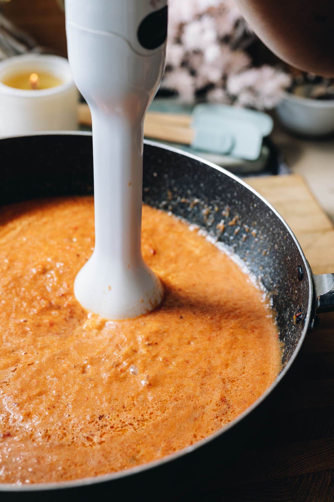 A large frying pan sits on a wooden board. Inside is soup that is being blended with a stick blender. 