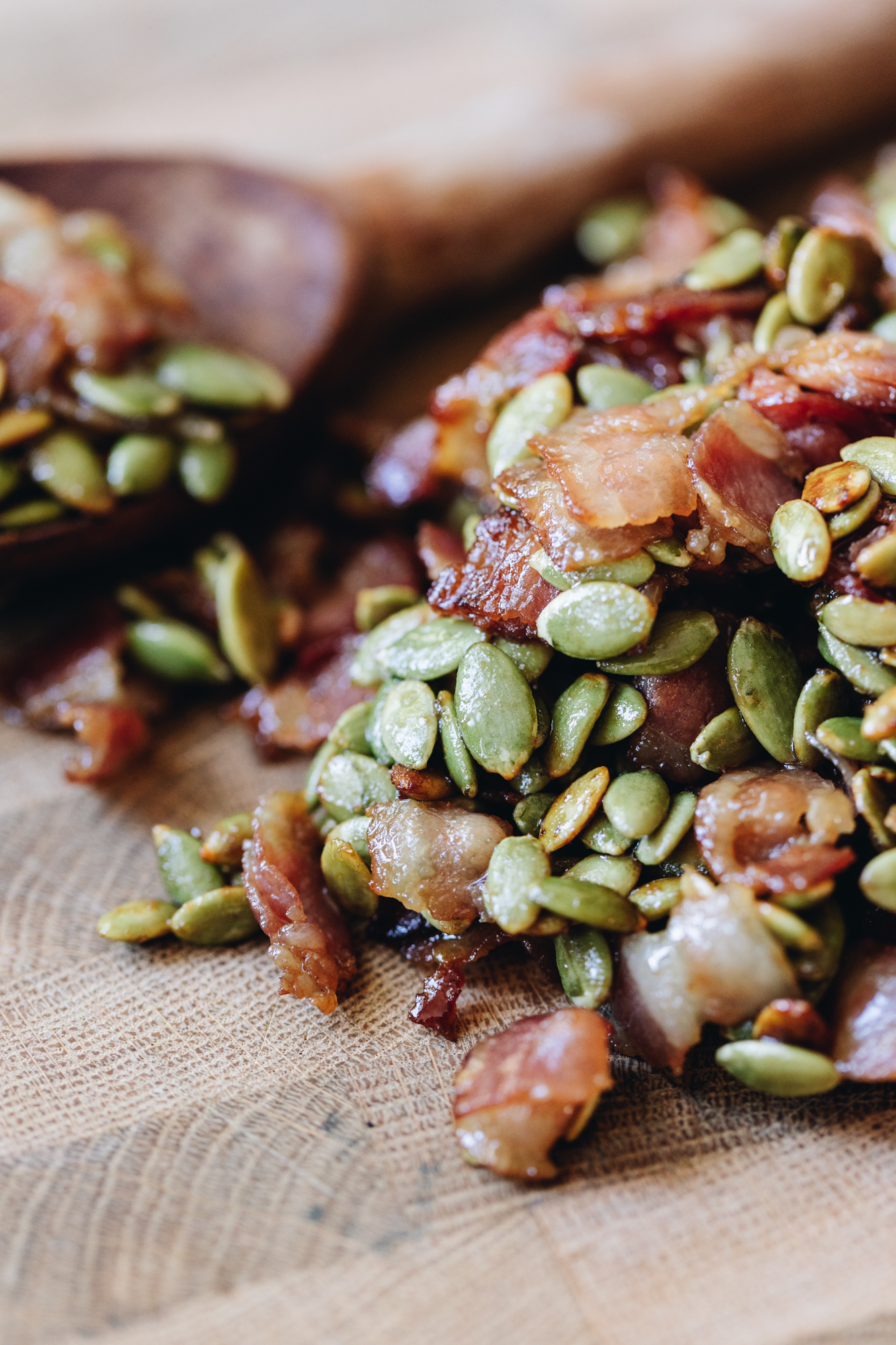 On a wooden board is a pile os sticky bacon and pumpkin seeds. A wooden spoon is in the background. 