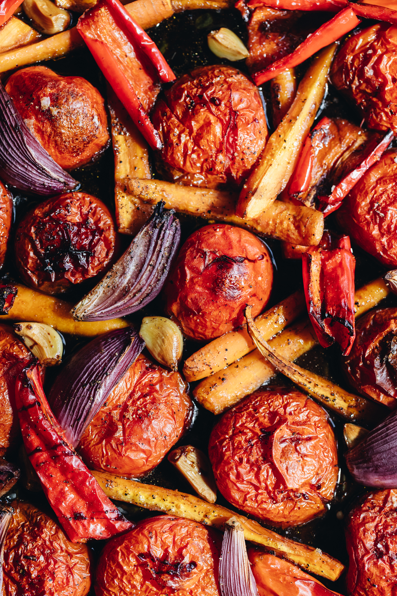 A roasting tray lined with baking paper sits on a stone bench. In the tray is tomatoes, carrots, onions, capsicums and garlic cloves drizzle with a balsamic glaze, they are all roasted.