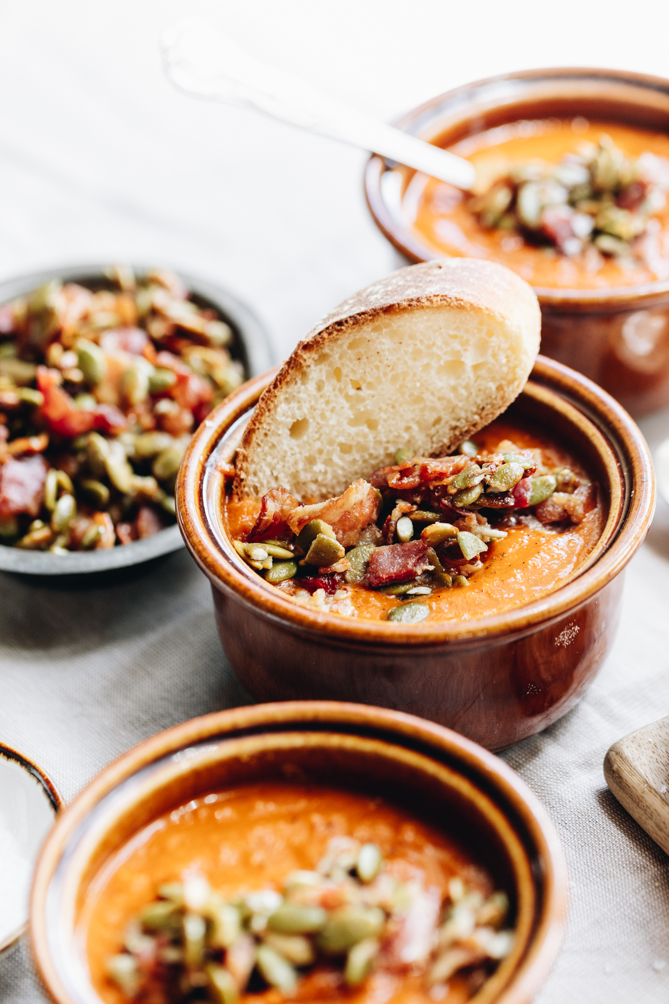 Three easy roasted tomato soup bowls are on a tablecloth. The soup is topped with bacon and pumpkin seeds and the centre one has a piece of baguette sticking out of it. 