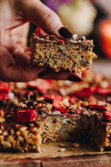 Peanut butter Rice Bubble Slice is on a wooden board on a brown baking paper. One piece is being held in a hand showing the texture.