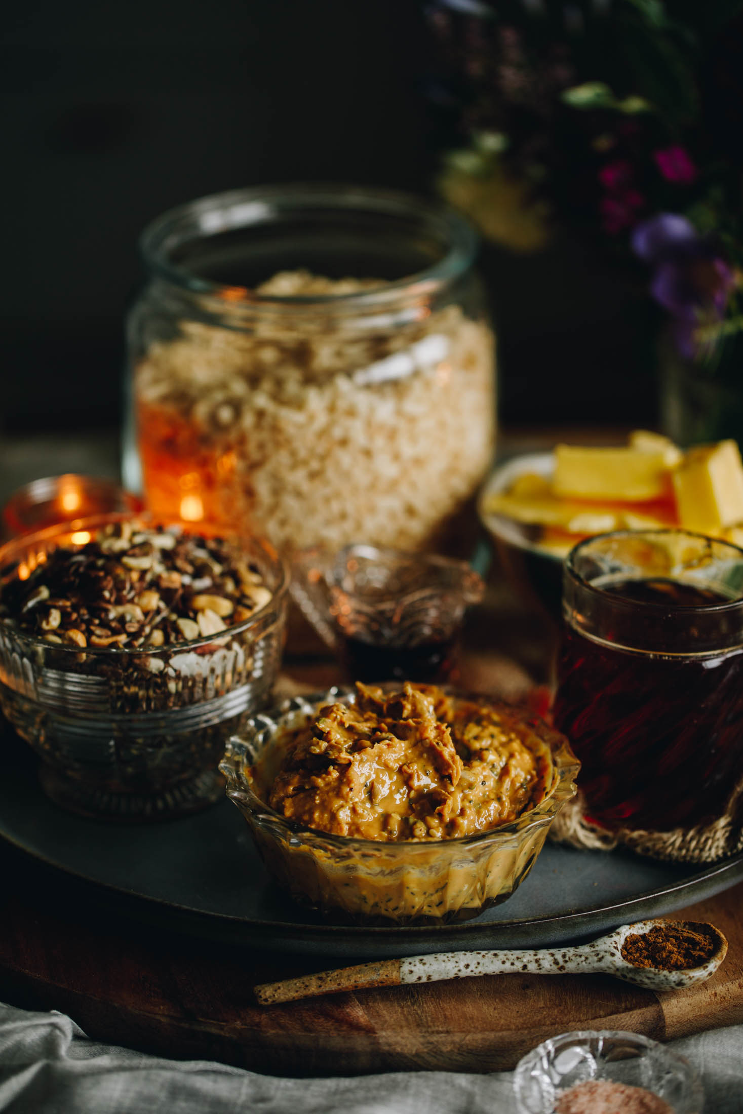 A wooden board sits on a cream cloth on a wooden table. On the board is a blue plate with ingredients on it in vintage bowls and jars. There is seeded peanut butter, rice syrup, keto muesli, butter and Rice Bubbles in view with flowers in the background. A ceramic spoon sits at the from of the planet with cinnamon in it. 
