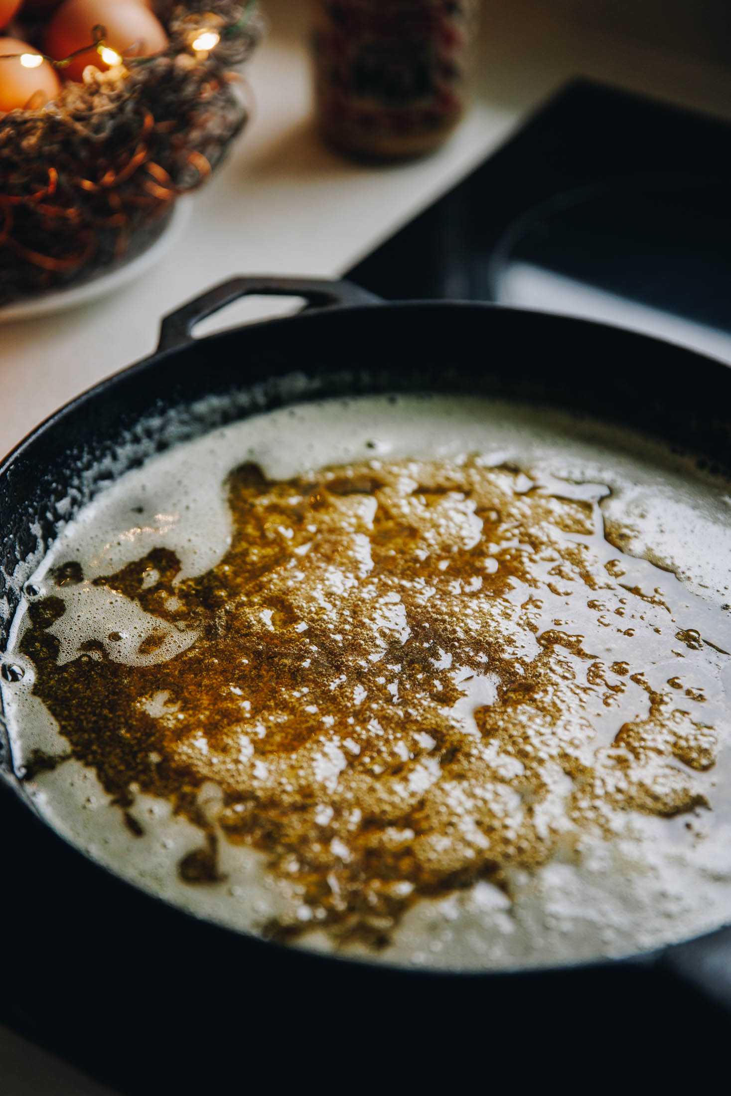 A black cast iron pan sits on a black stove top. In it is a simple caramel bubbling. Eggs in a wire basket are in the background. 