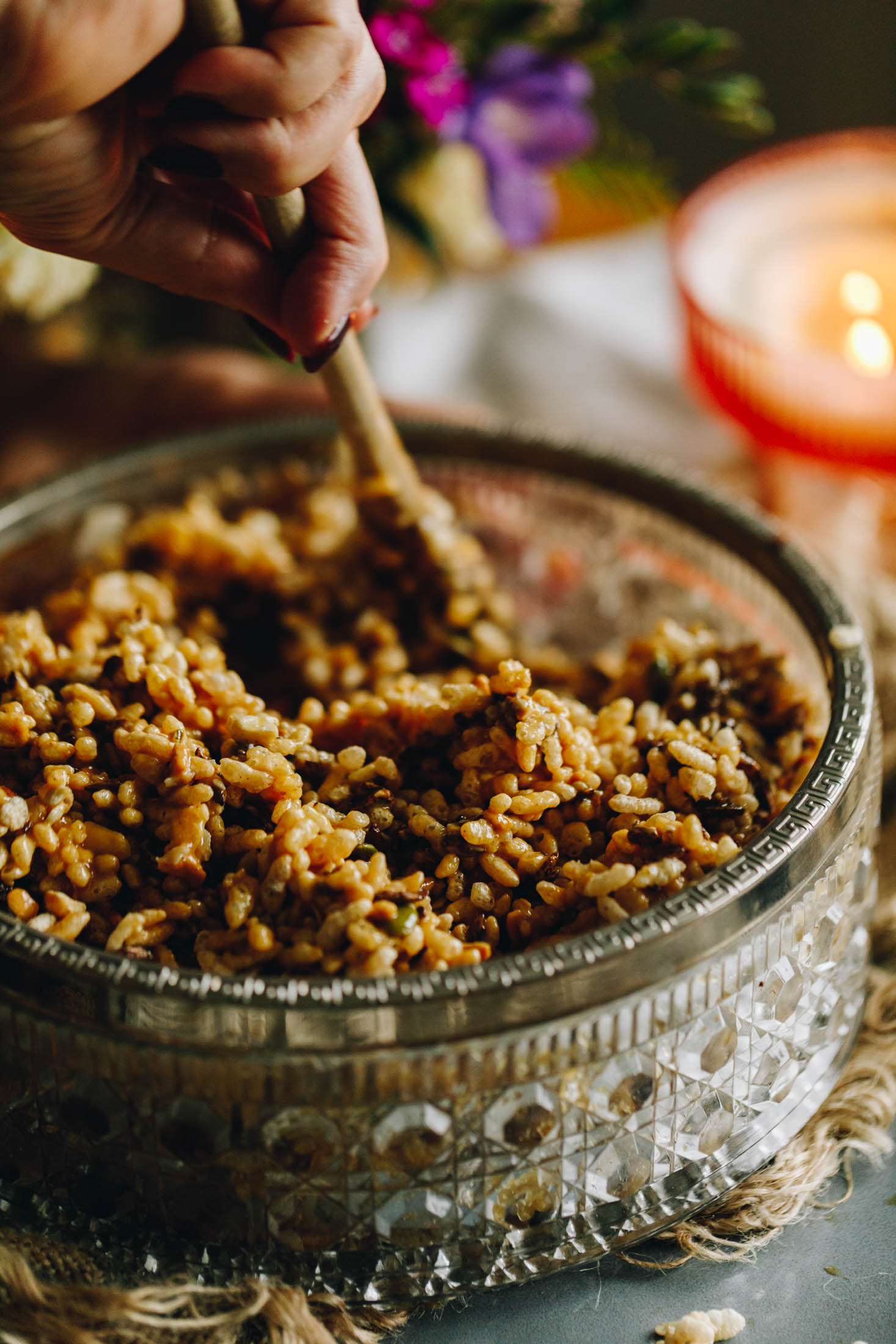 A vintage bowl sits on a blue plate with natural fabric under it. In the bowl is a Rice Bubble mixture that is being stirred with a wooden spoon. A orange candle is burning in the background with purple flowers. 