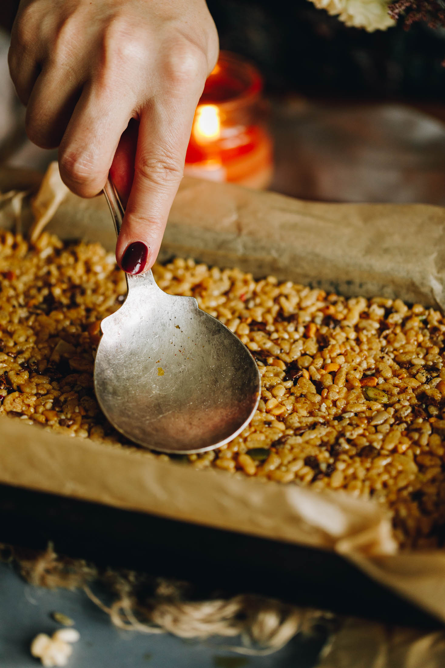 A black tin lined with brown baking paper sits on natural fabric. In the tin is Peanut Butter Rice Bubble Slice that is being pressed down firmly with a large vintage spoon. 