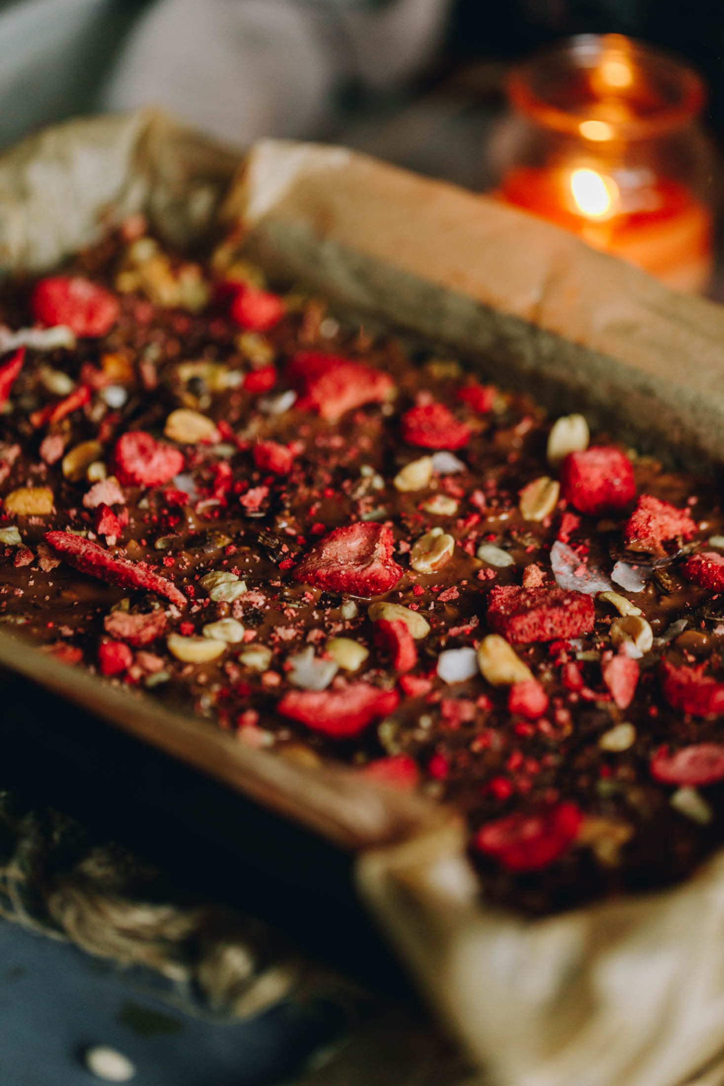 A black tin lined with brown baking paper sits on natural fabric. In the tin is Peanut Butter Rice Bubble Slice that has been decorated with chocolate, keto muesli and freeze-dried strawberries.