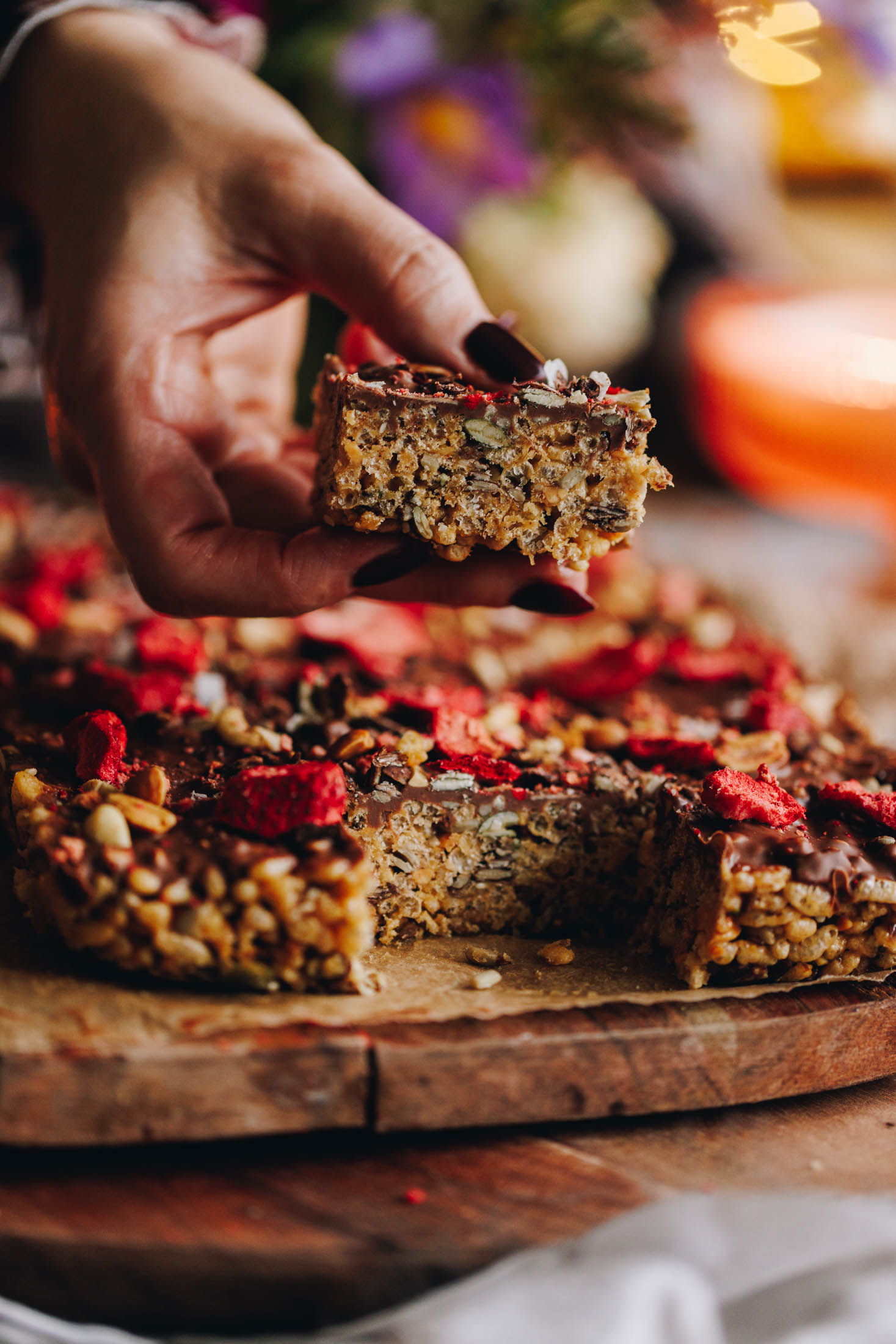Peanut butter Rice Bubble Slice is on a wooden board on a brown baking paper. One piece is being held in a hand showing the texture and the chocolate and freeze-dried strawberry topping. 