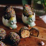 Peanut Butter Chocolate Eggs are on a wooden board. They are covered in chocolate and sprinkles. Three are sitting on the left edge of frame. One is cut open showing the peanut butter and marshmallow filling. Two of the eggs are in small chicken egg cups.