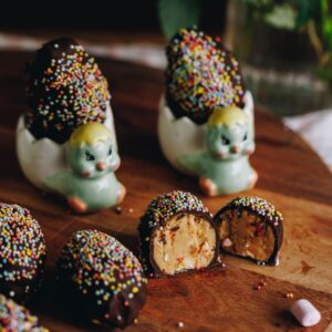 Peanut Butter Chocolate Eggs are on a wooden board. They are covered in chocolate and sprinkles. Three are sitting on the left edge of frame. One is cut open showing the peanut butter and marshmallow filling. Two of the eggs are in small chicken egg cups.