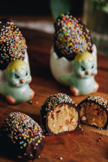 Peanut Butter Chocolate Eggs are on a wooden board. They are covered in chocolate and sprinkles. Three are sitting on the left edge of frame. One is cut open showing the peanut butter and marshmallow filling. Two of the eggs are in small chicken egg cups.