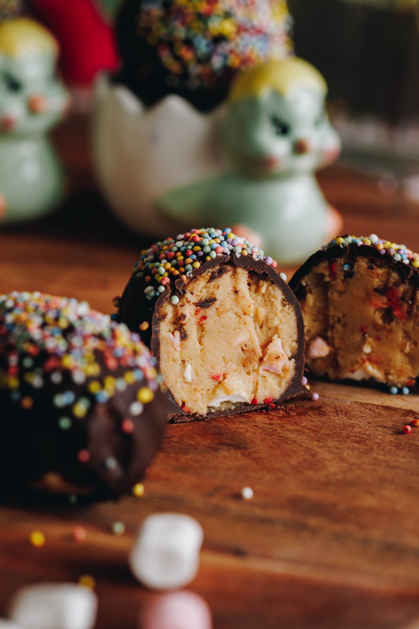 Peanut Butter Chocolate Eggs are on a wooden board. They are covered in chocolate and sprinkles. One is on the left edge of frame. One is cut open showing the peanut butter and marshmallow filling. Two of the eggs are in small chicken egg cups in the background.
