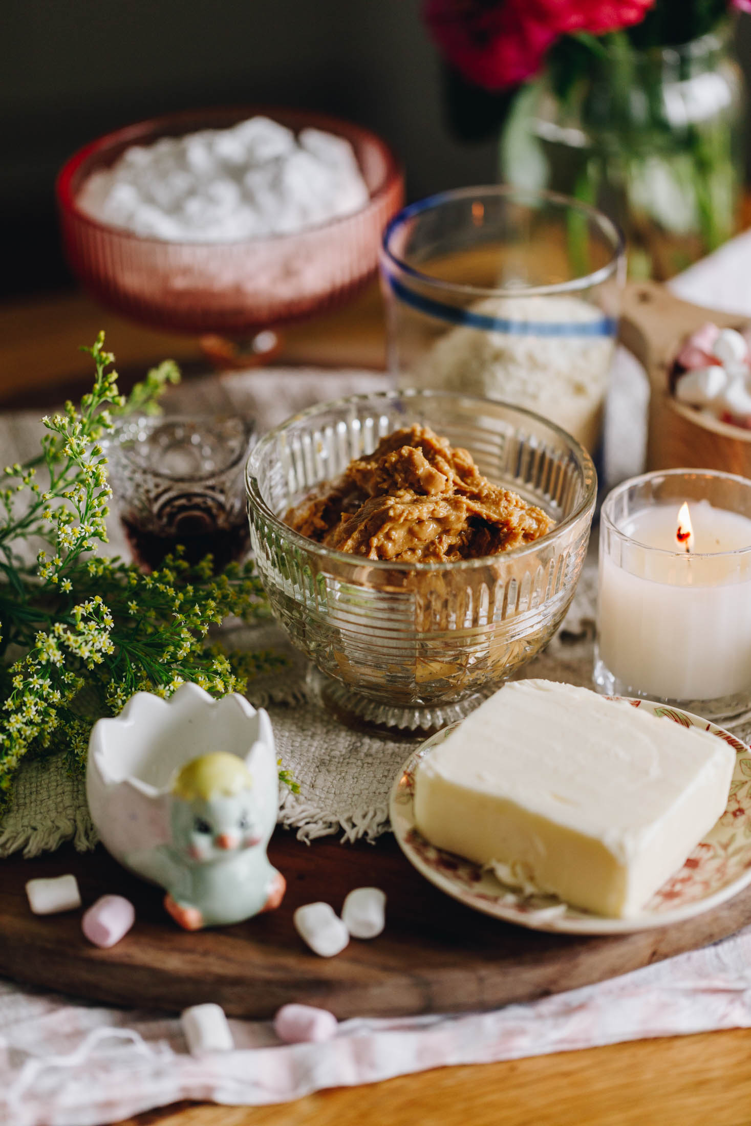 A wooden board sits on a white cloth on a wooden table. ON the board is a chicken egg cup and greenery. Next to it is peanut butter, butter, a white candle, ground almonds and icing sugar in vintage cups. Pink flowers are in the background.