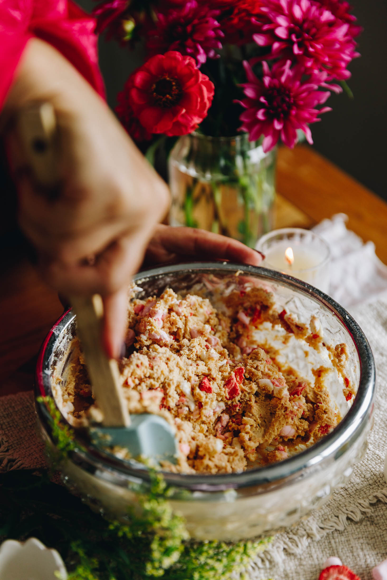 A vintage glass bowl sits on a cream cloth, on a wooden table. In the bowl is butter mix with chopped marshmallows and free-dried strawberries. It is being mixed with a wooden spatula with a blue handle. Behind the bowl is pink flowers in a vase.