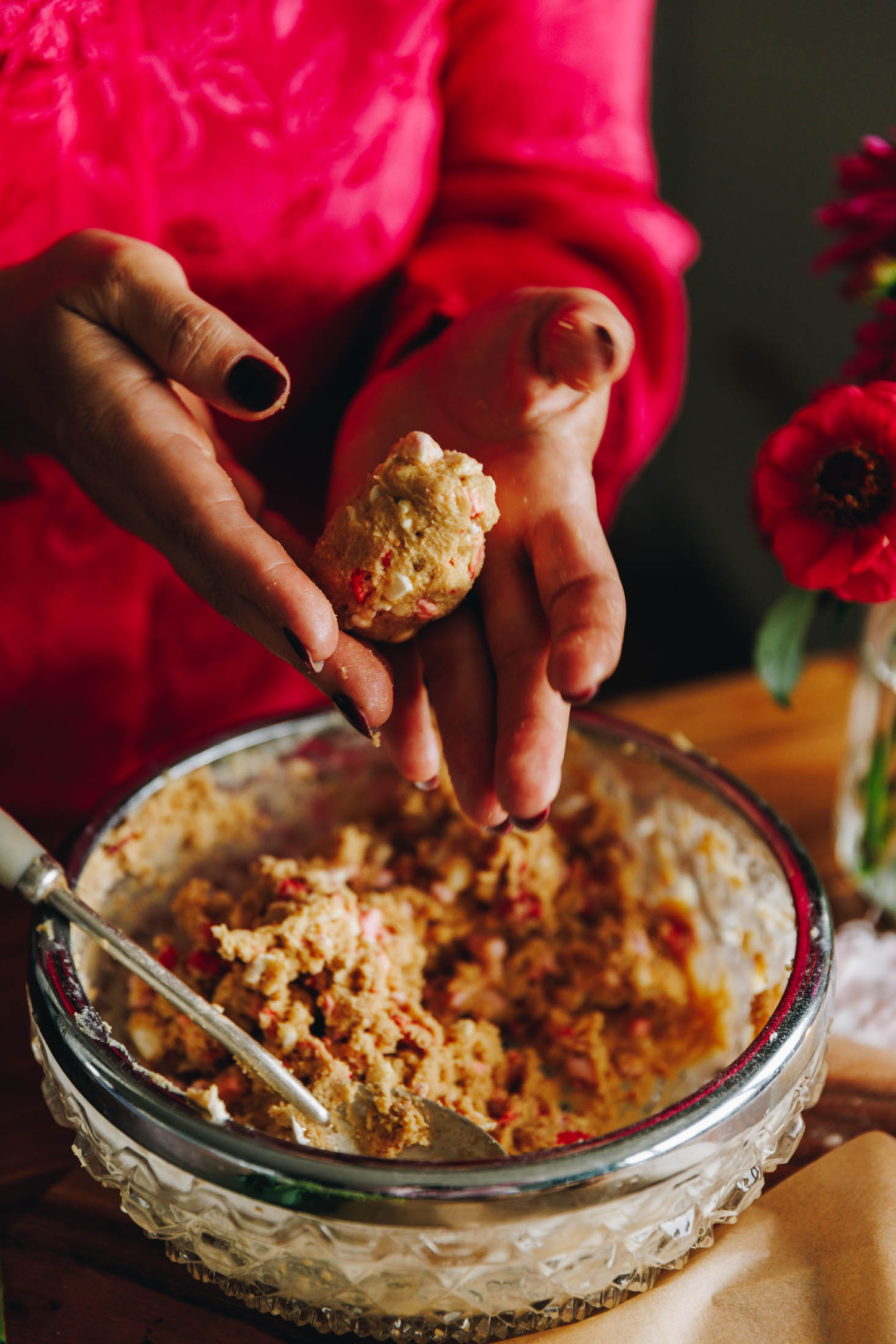 A vintage glass bowl sits on a cream cloth, on a wooden table. In the bowl a peanut butter easter egg mix with chopped marshmallows and free-dried strawberries. Naomi is moulding one in to an egg shape with her hands hovering above the bowl. Behind the bowl is pink flowers in a vase.