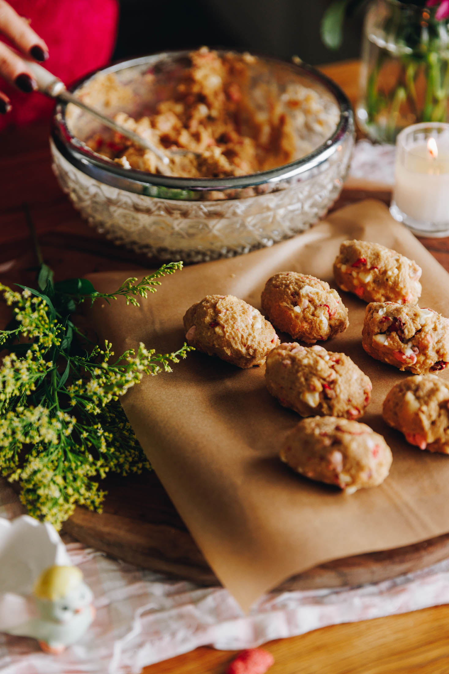 A wooden board sirs on a cream cloth on a wooden table. On the board is a rectangle board lined with brown baking paper. It is rolled peanut butter easter eggs with chunks of mini marshmallows and freeze-dried strawberries. In the background is the bowl with remaining mixture. Greenery is next to the wooden board.