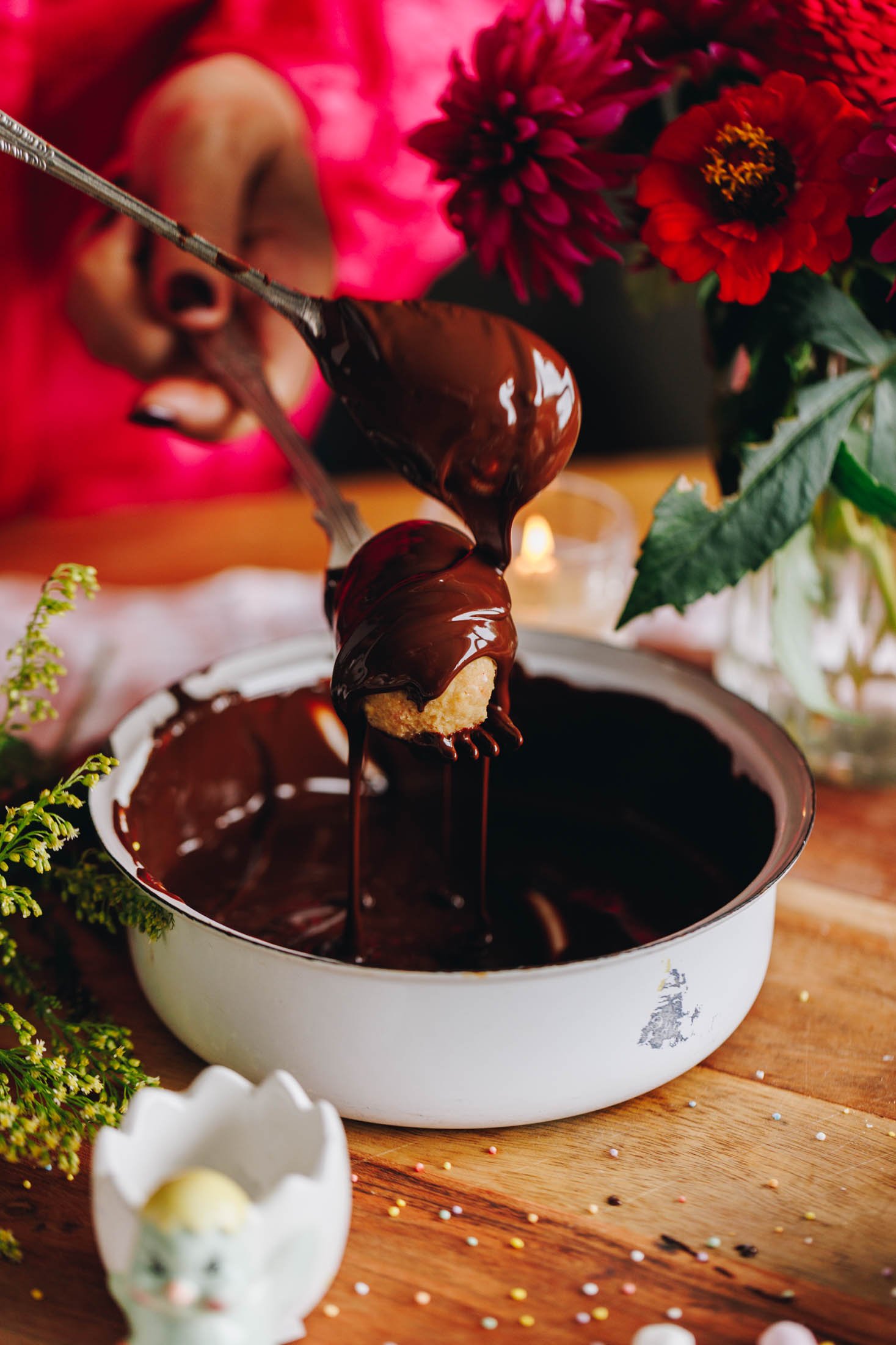 A white metal bowl sits on a wooden board. In the bowl is melted chocolate. Naomi is using a spoon to drizzle chocolate all over a peanut butter egg. The egg is sitting on a fork. A bird egg cup is on the board as well and pink flowers are in the background.
