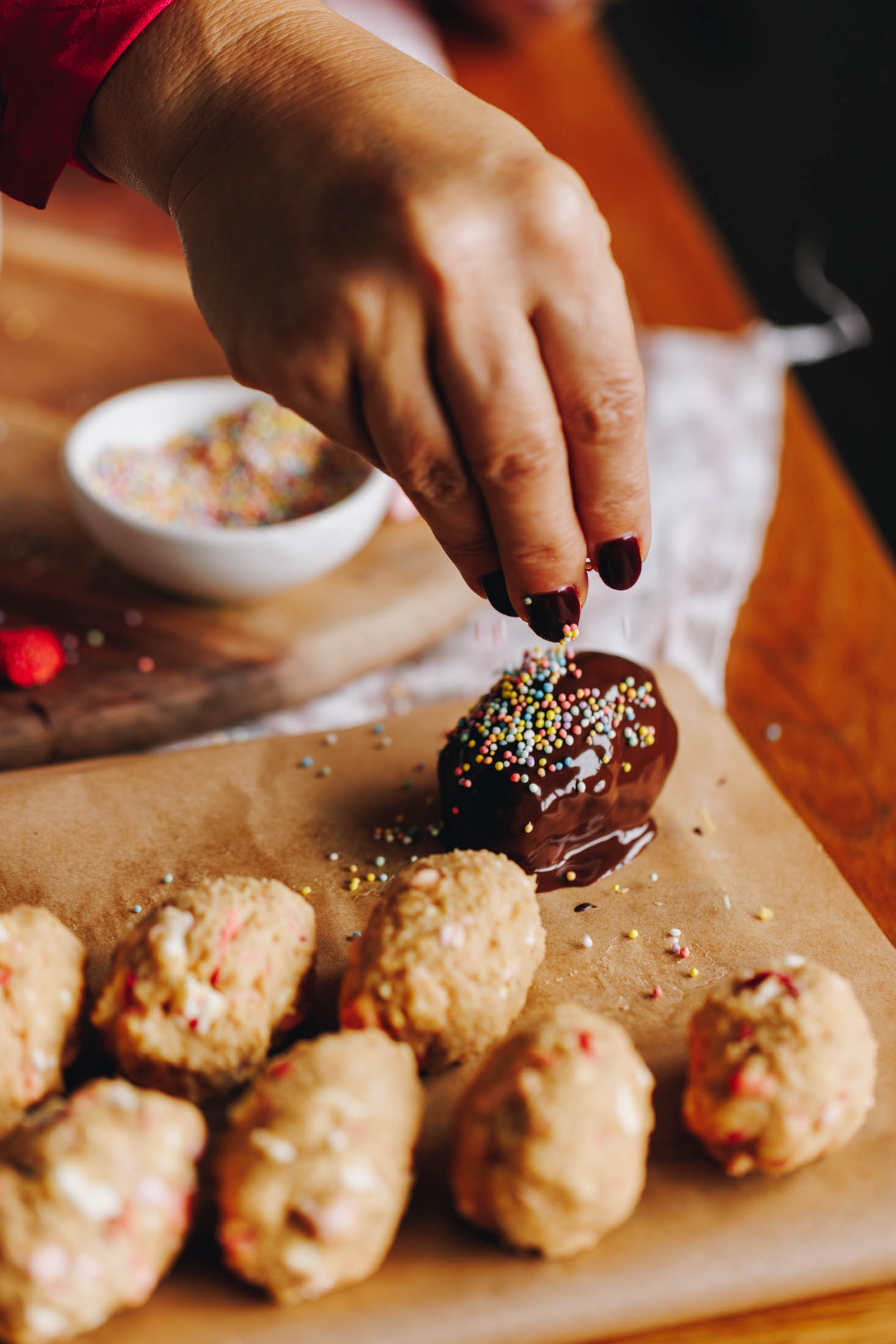 A wooden board lined with baking paper sits on a wooden table. On the paper is easter eggs. One has been dipped in chocolate and is being covered in sprinkles. The others are uncovered.