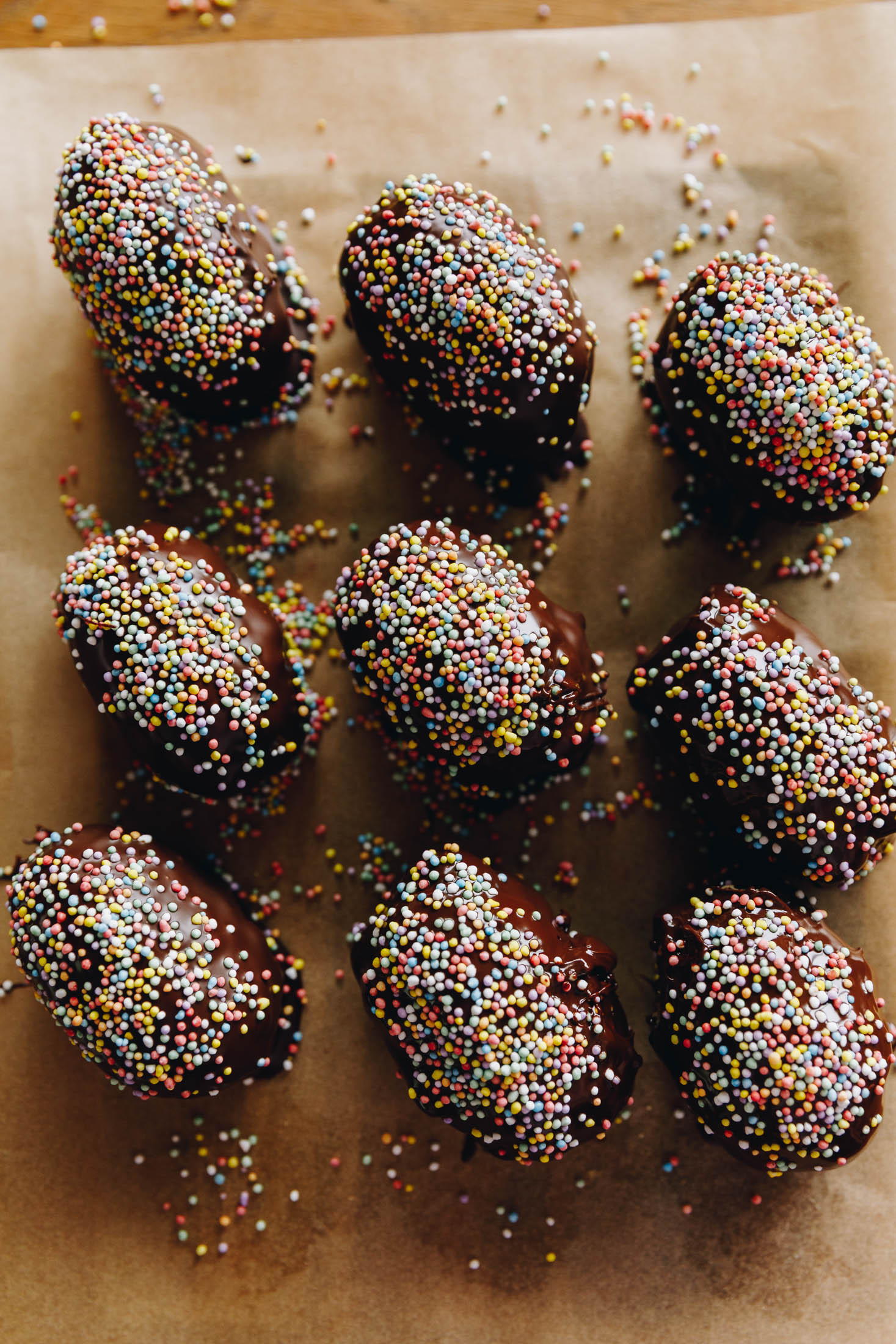 A flat lay shot shows 9 peanut butter easter eggs lined up on a wooden board lined with baking paper. They are covered in chocolate and sprinkles.