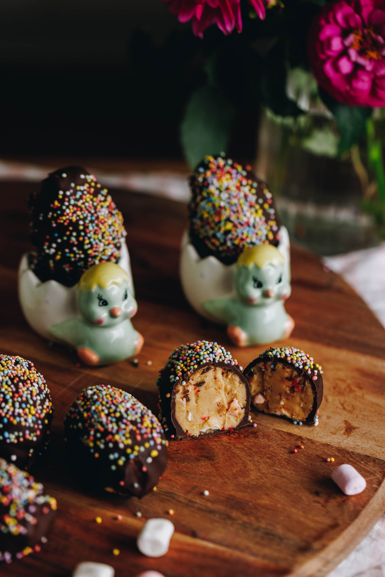 Peanut Butter Chocolate Eggs are on a wooden board. They are covered in chocolate and sprinkles. Three are sitting on the left edge of frame. One is cut open showing the peanut butter and marshmallow filling. Two of the eggs are in small chicken egg cups.