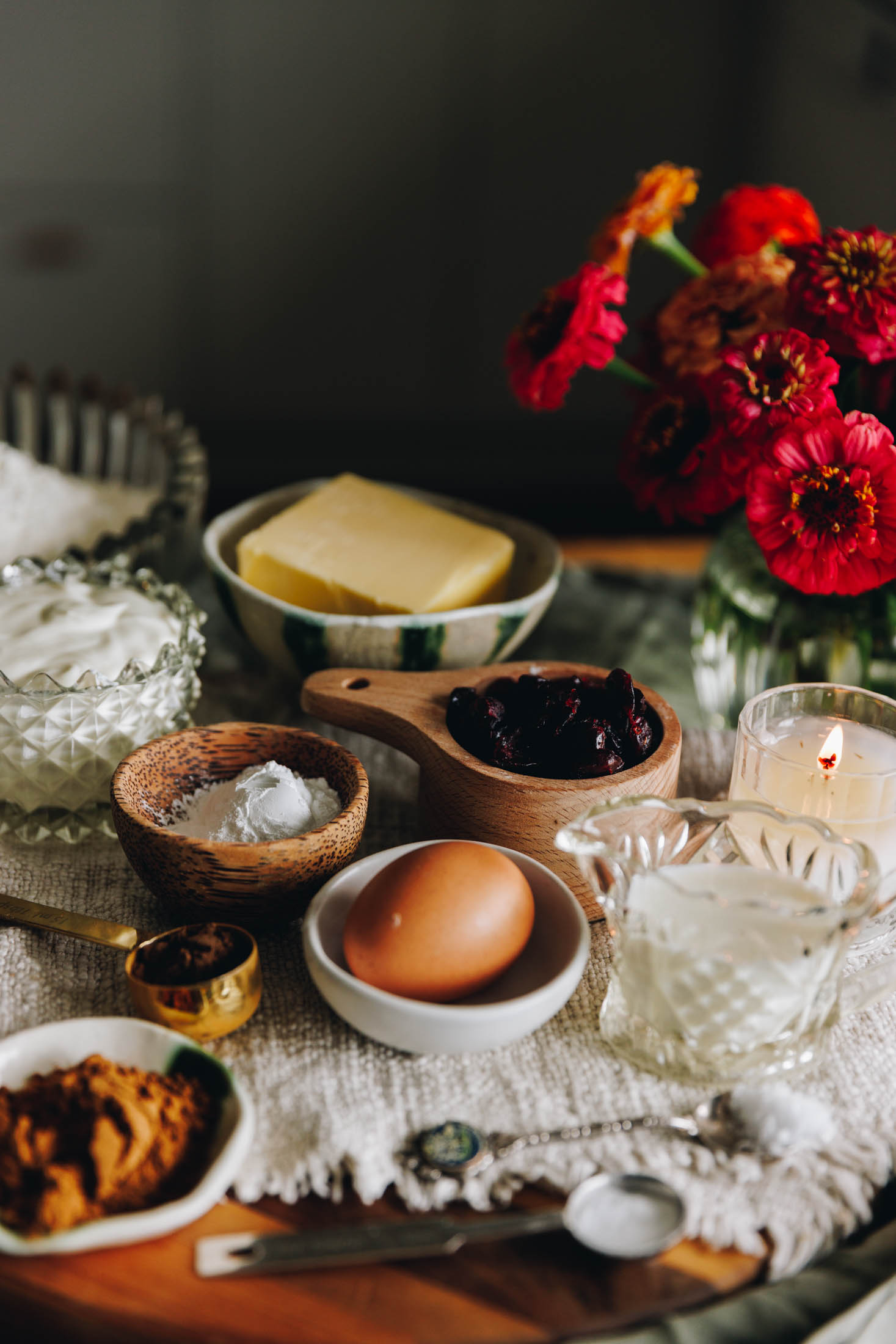 A wooden board sits on a green table cloth. On the board is a cream cloth. On top of the cloth is vintage bowls and jars with ingredients in them. There are spices, an egg, milk, baking powder, cranberries, butter, yogurt, salt and flour in view. On the right hand side is a glass vase with red and pink flowers in it.