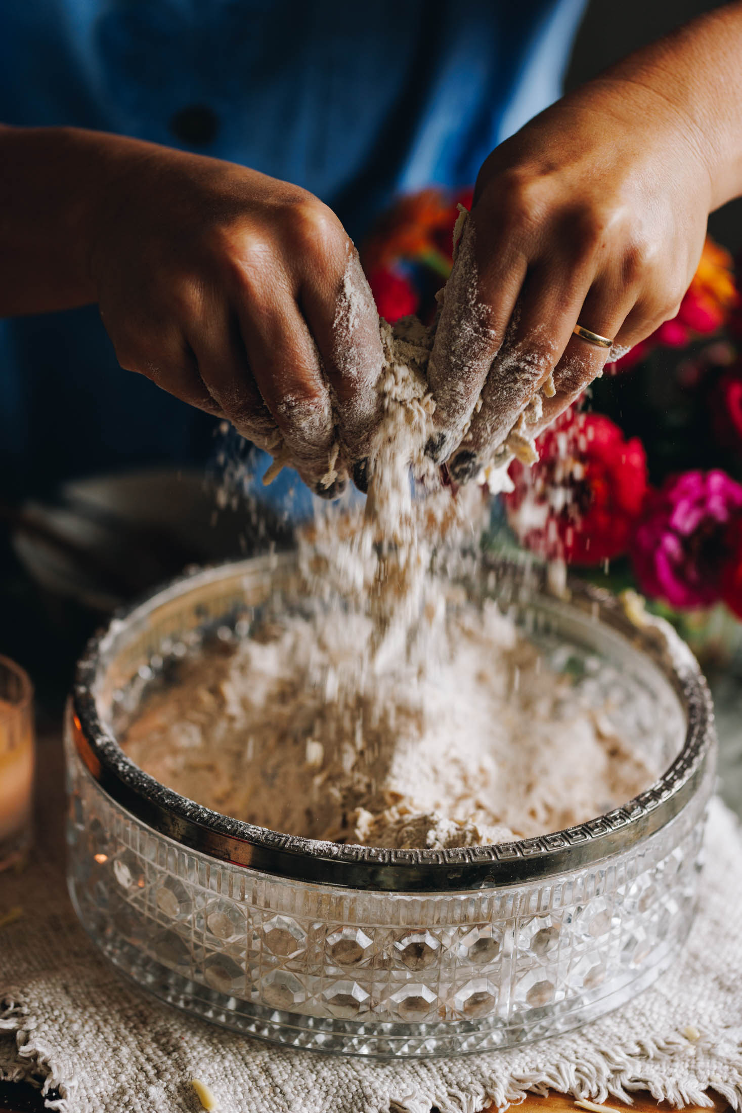 A glass vintage bowl sits on a cream cloth on a wooden table. In the bowl is a flour mixture that is being lifted out of the bowl by Naomi's hands as she rubs the butter in to the dry ingredients. Pink and orange flowers are in the background.