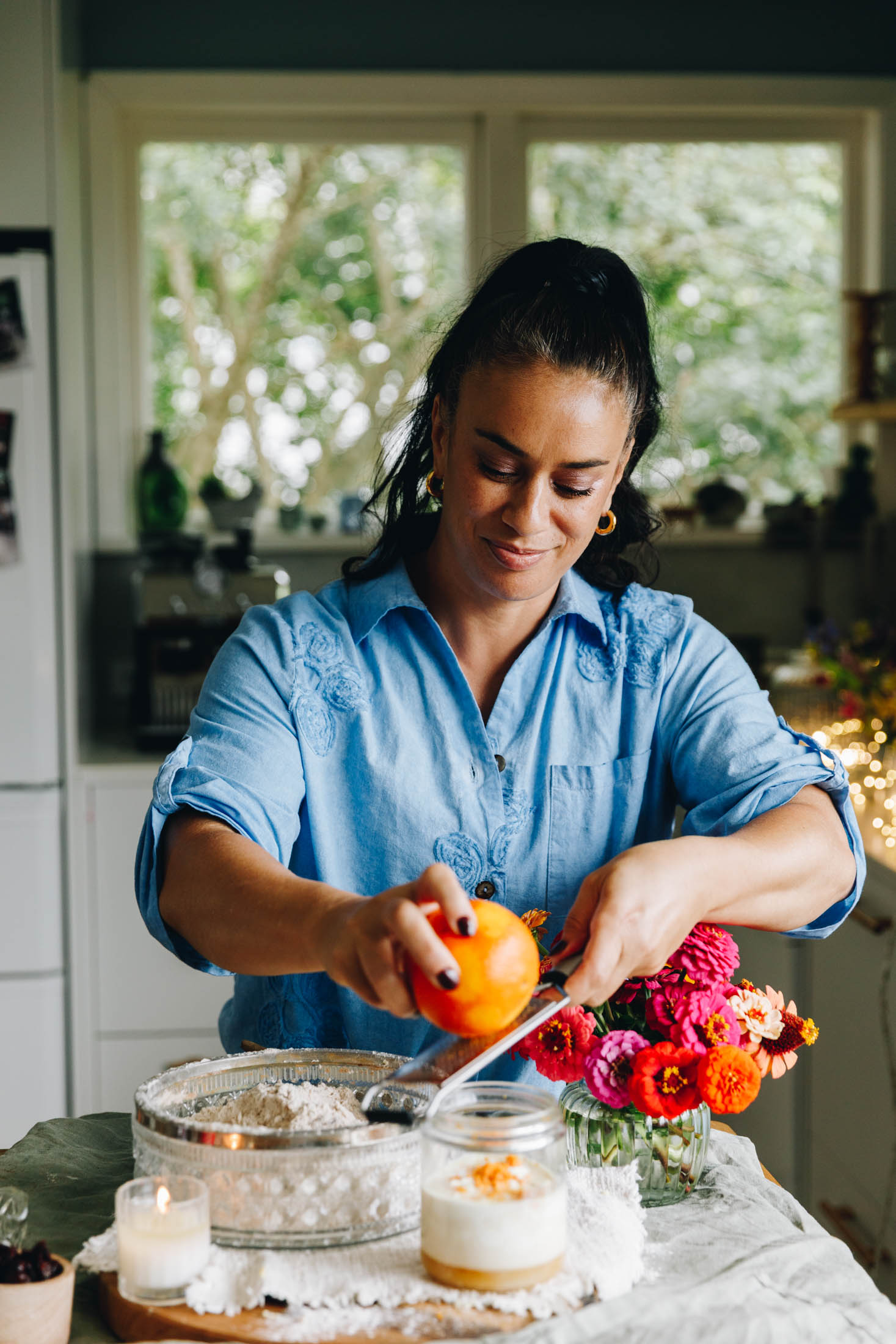 Naomi Toilalo is finely grating orange zest in to a glass jar in her kitchen. There is a flour mix in a bowl next to there with orange and pink flowers. The table has a cream tablecloth on it. Fairy lights are in the background.