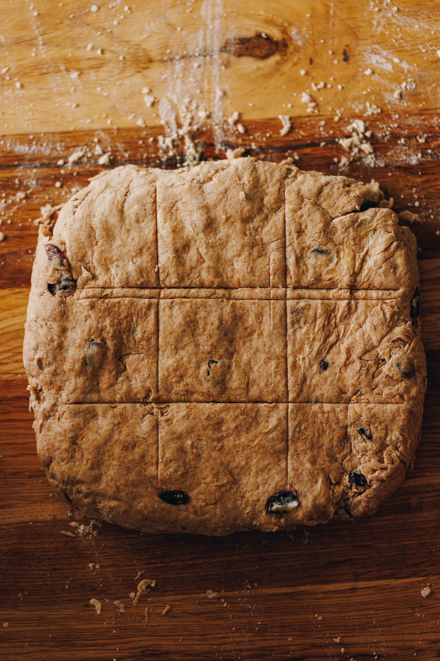 A spiced dough sits on a wooden table that has been dusted with flour. The dough is in a square shape and has lines across it, diving it in to 9 pieces.