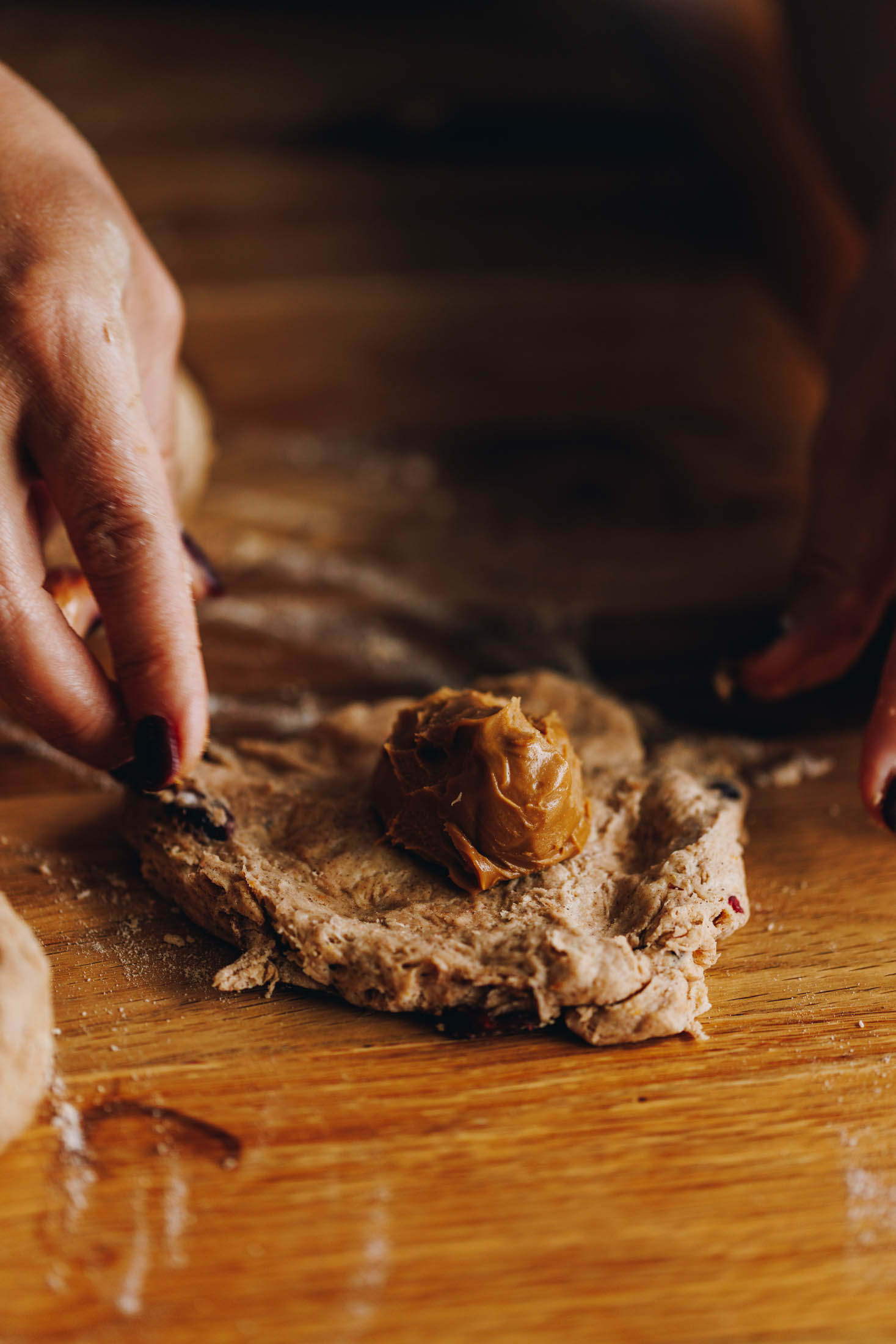 A square piece of scone dough sits on a wooden table. The dough has been pressed out and a tablespoon of biscuit spread sits in the centre. Naomi has hold of the corners, ready to fold them in.