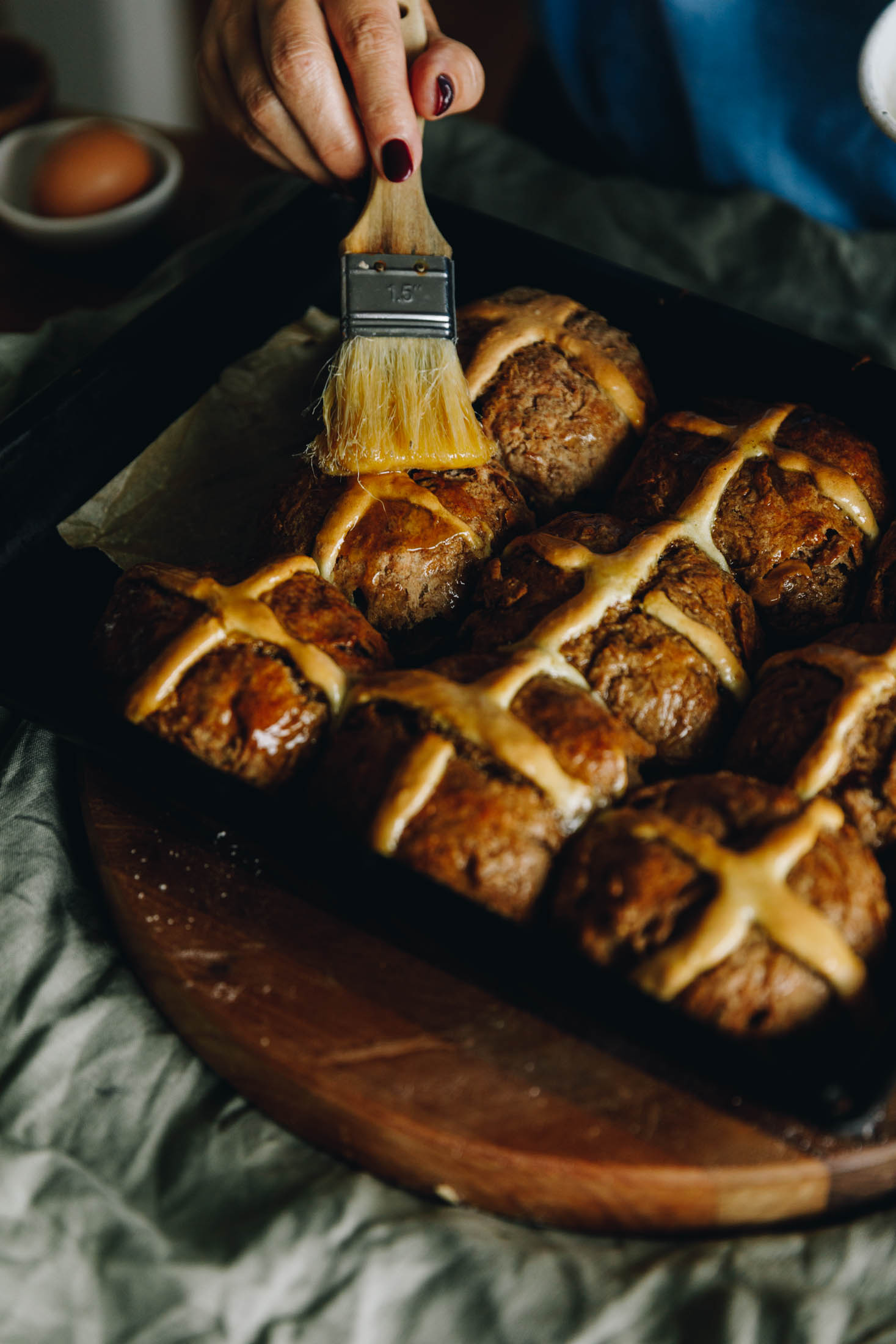 Hot Cross Buns are being glazed in a black braking tray. The tray is sitting on a round wooden board, on a green table cloth.