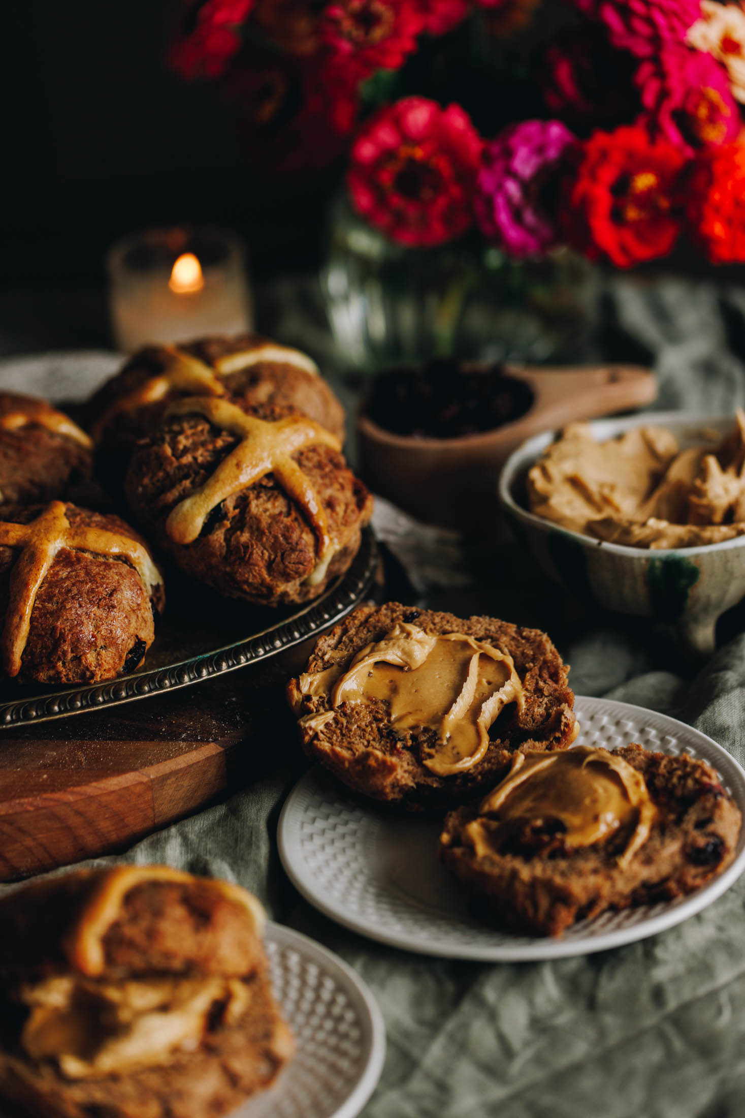 A green tablecloth is on a table. On the cloth is a small white plate with hot cross bun scones spread with a biscuit spread butter. A silver tray is behind the plate with whole hot cross bun scones on it. Next to that is a ceramic bowl with whipped biscuit spread and a wooden cup with cranberries.